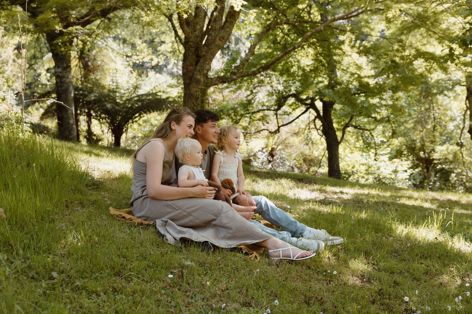 Family sitting together on a grassy hillside under shady trees on a sunny summer afternoon. Tauranga family photoshoot