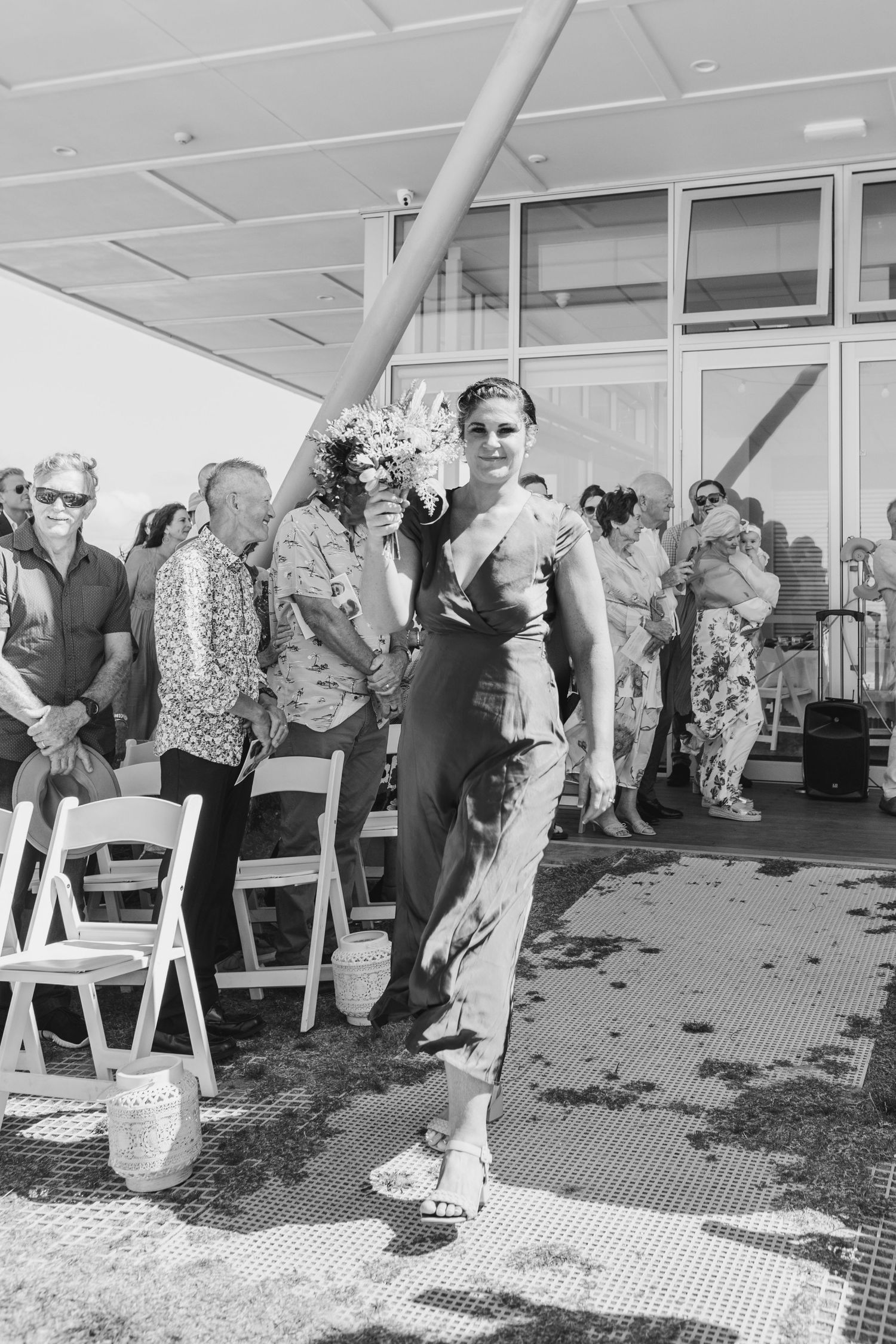 Wedding guests watch as attendees walk down the aisle at an outdoor venue. Papamoa Surf Lifesaving Club Wedding