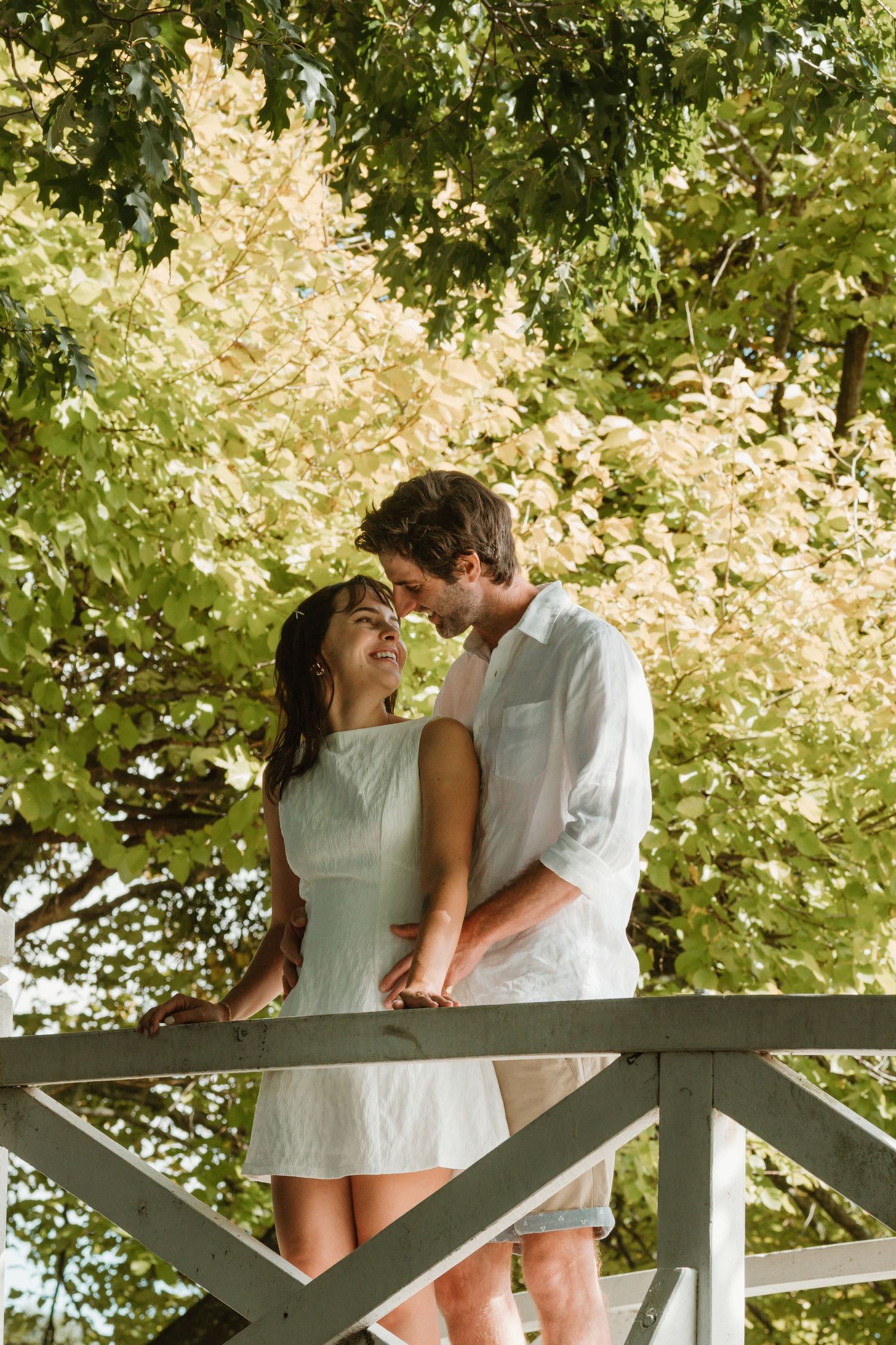 Romantic couple in white outfits sharing a tender moment on a wooden bridge surrounded by lush green trees.