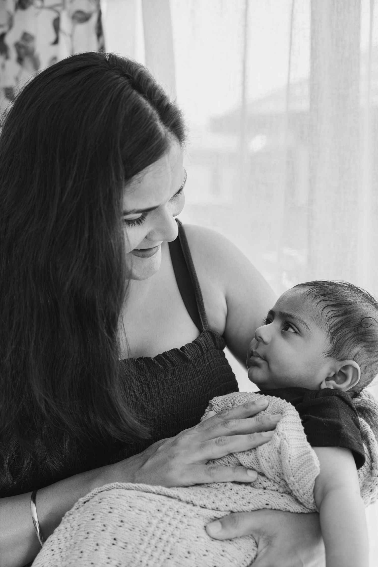 Mother in black dress lovingly holding and gazing at her infant baby wrapped in knitted blanket in black and white photo.