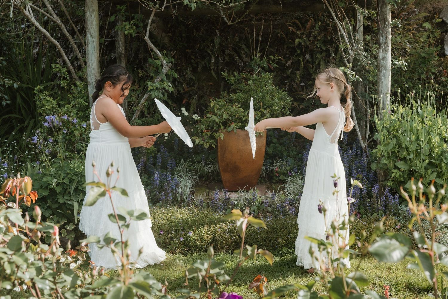 Two people in white dresses walk through a lush garden filled with blooming flowers and greenery.
