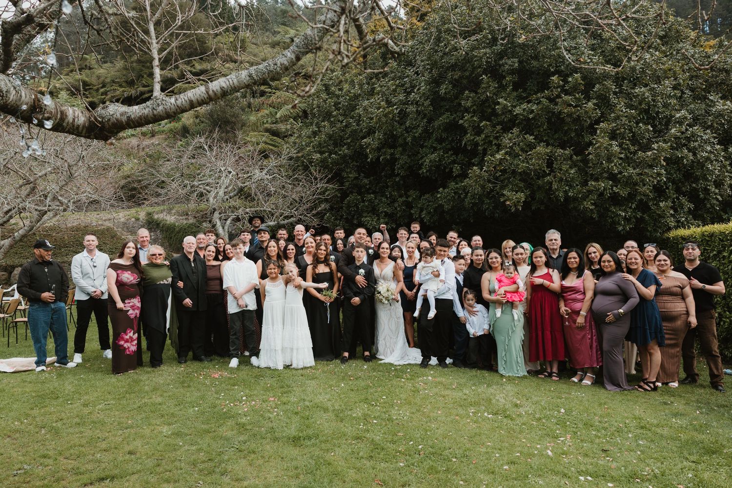 Large group of wedding guests pose together for panoramic photo on grassy lawn with trees in background.