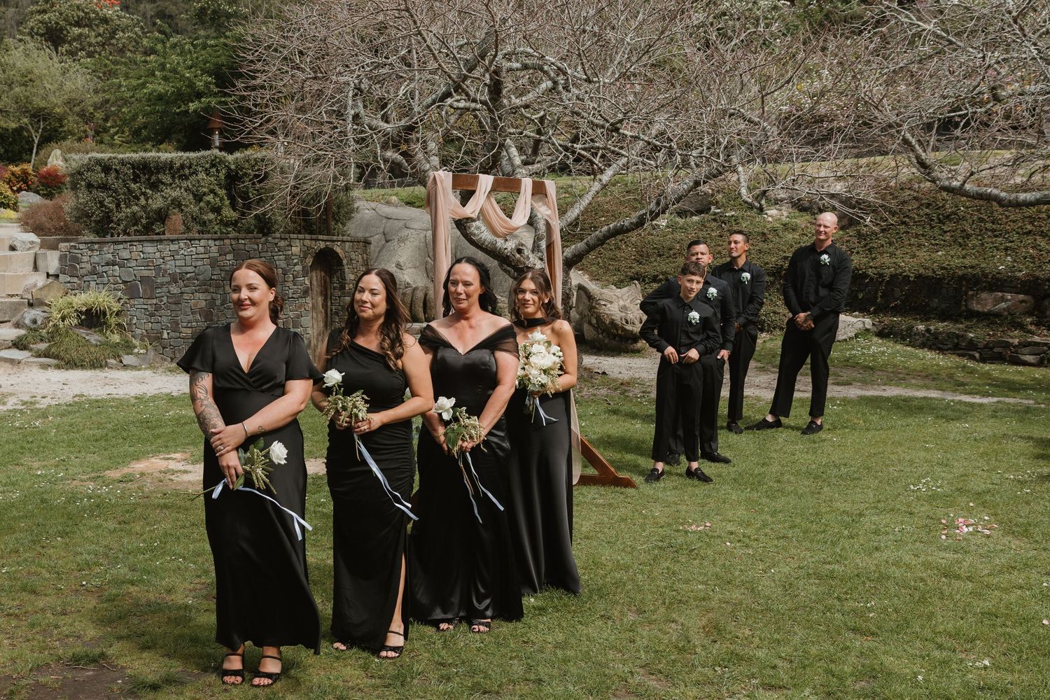 Wedding party in black formal attire poses together on grassy lawn during outdoor celebration.