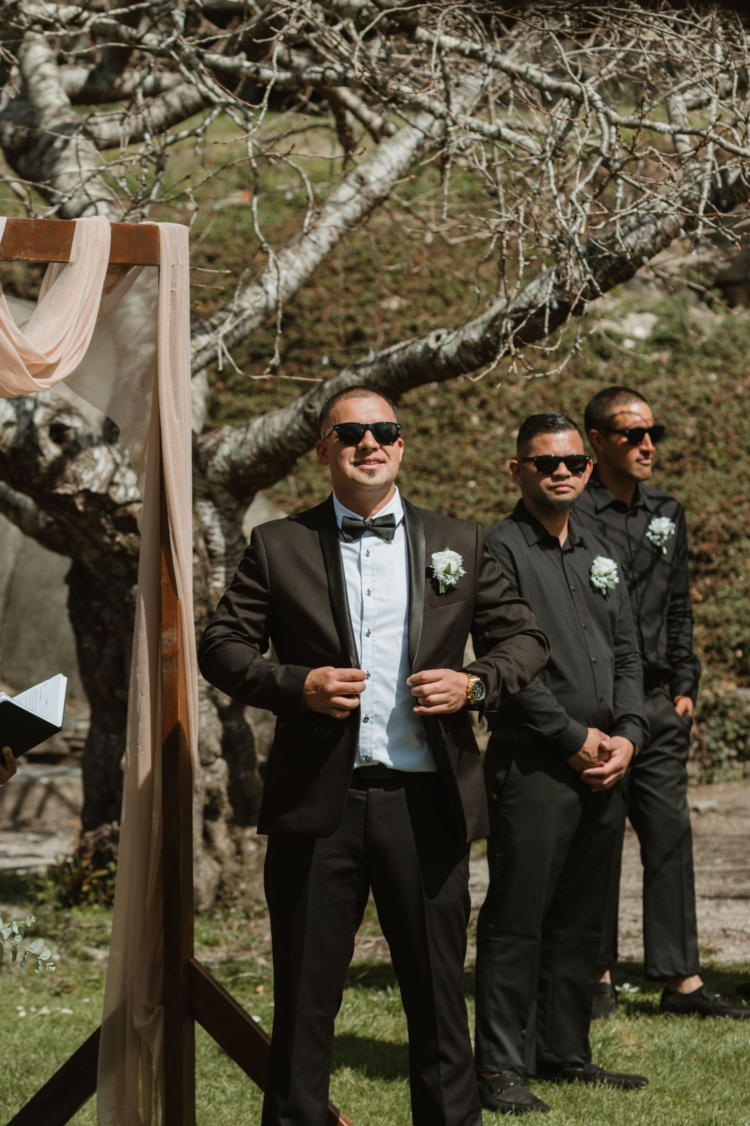 Groomsmen in black tuxedos and sunglasses stand casually outdoors near wooden posts during a wedding ceremony.