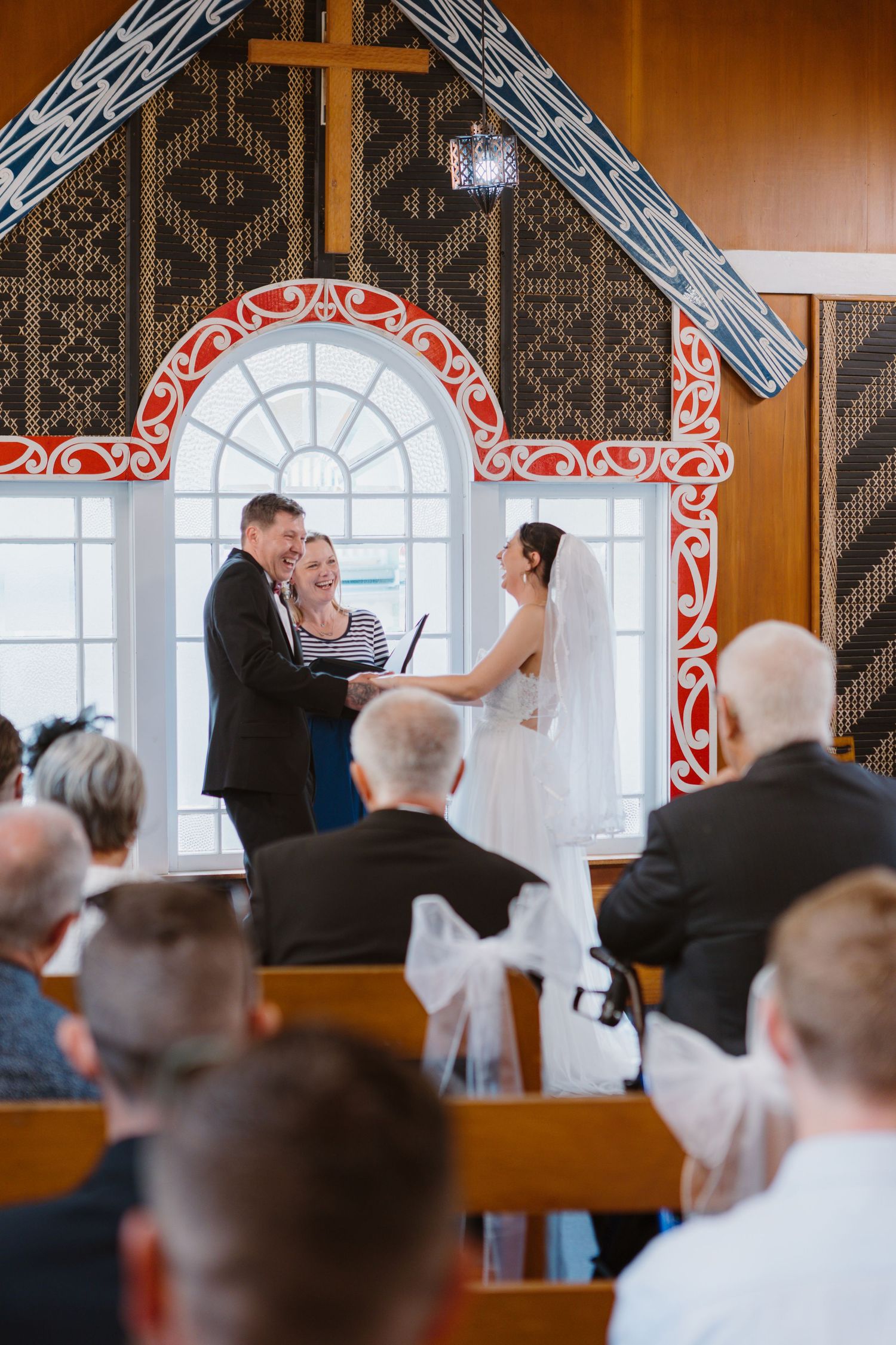 A wedding ceremony takes place inside a chapel with ornate red and white decorative trim around a large arched window.