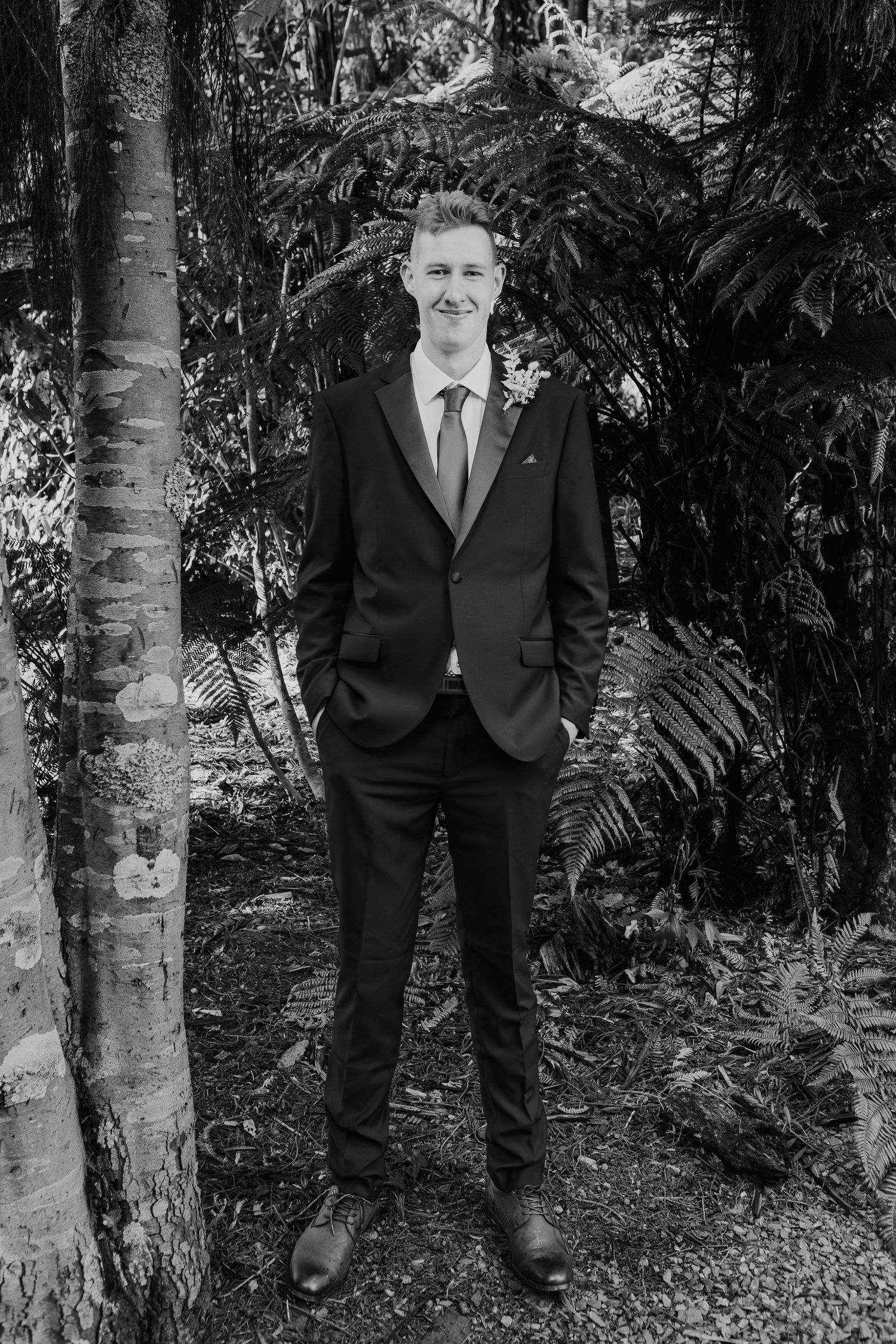 A person wearing a dark suit stands thoughtfully among trees and foliage in a black and white photograph.