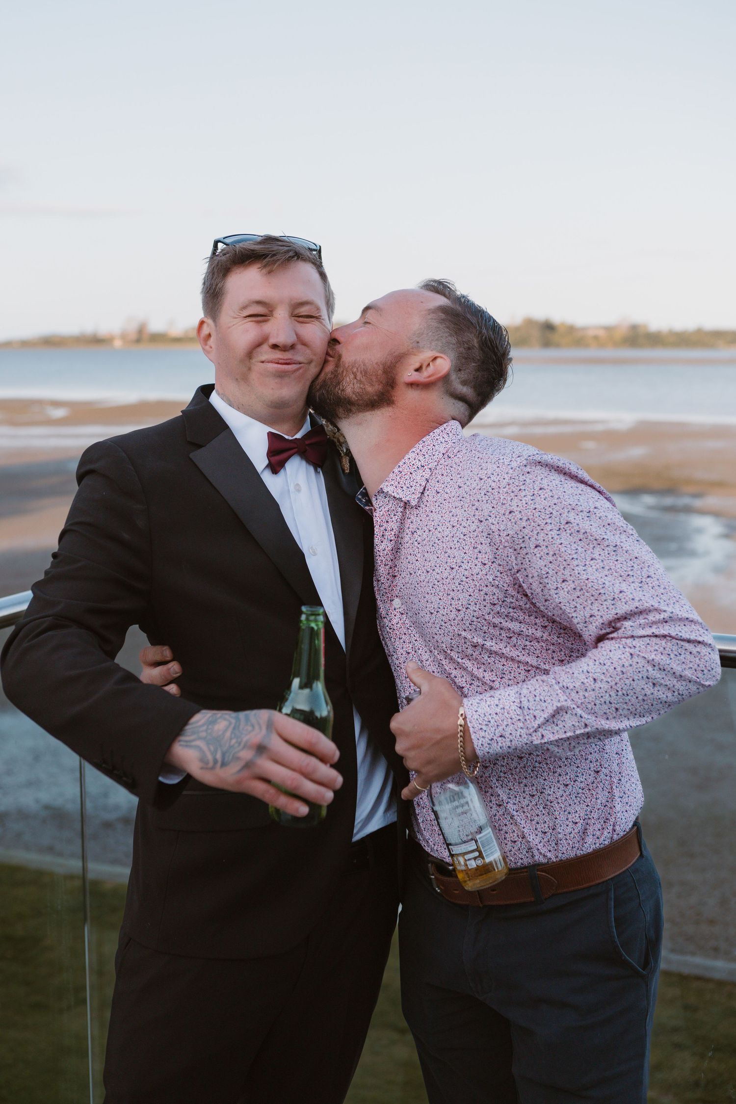 Two people in formal attire share a candid playful moment while holding drinks outdoors at sunset near the water.