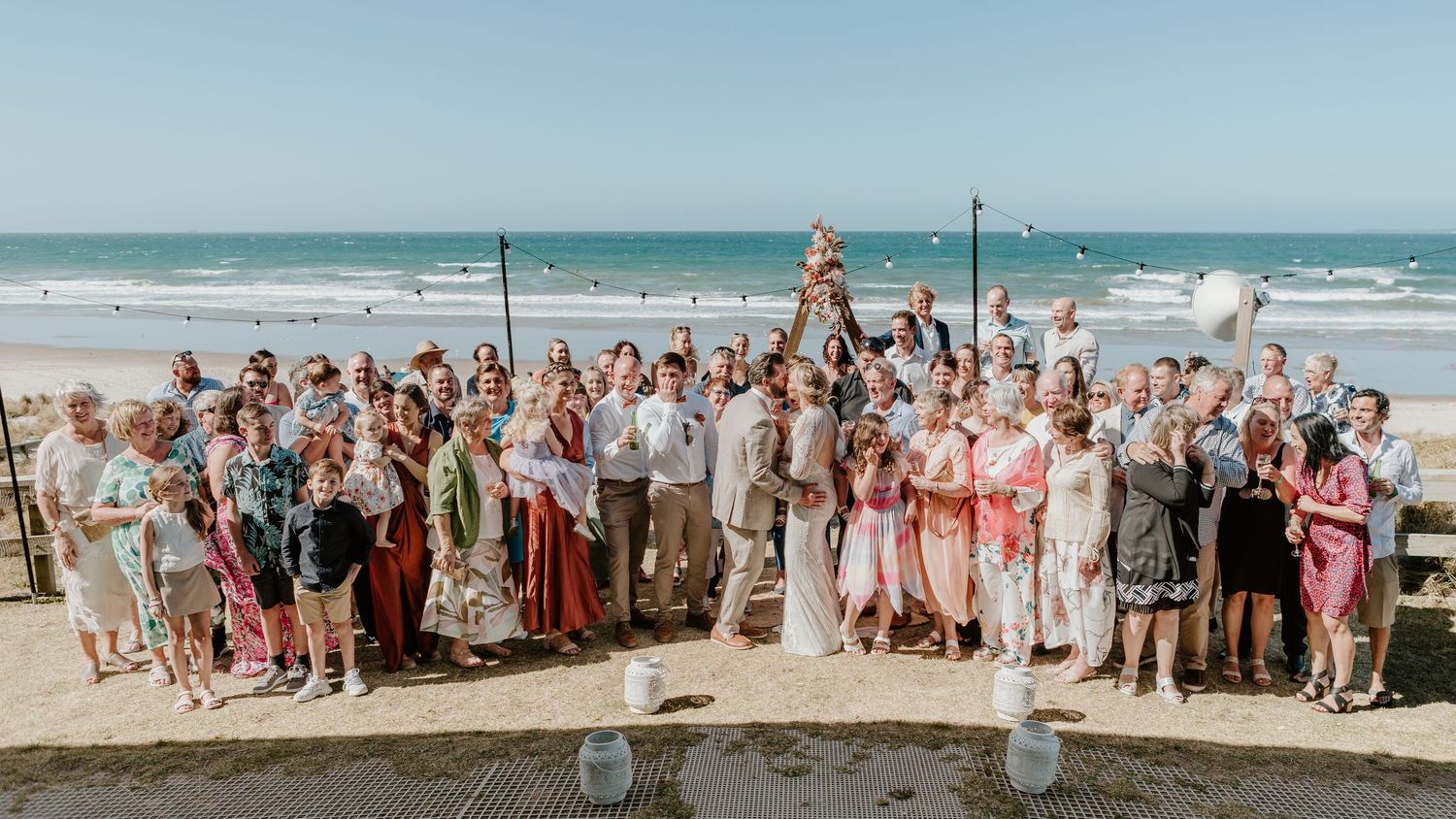A panoramic collage of wedding guests gathered together on a sunny beach for group photos.