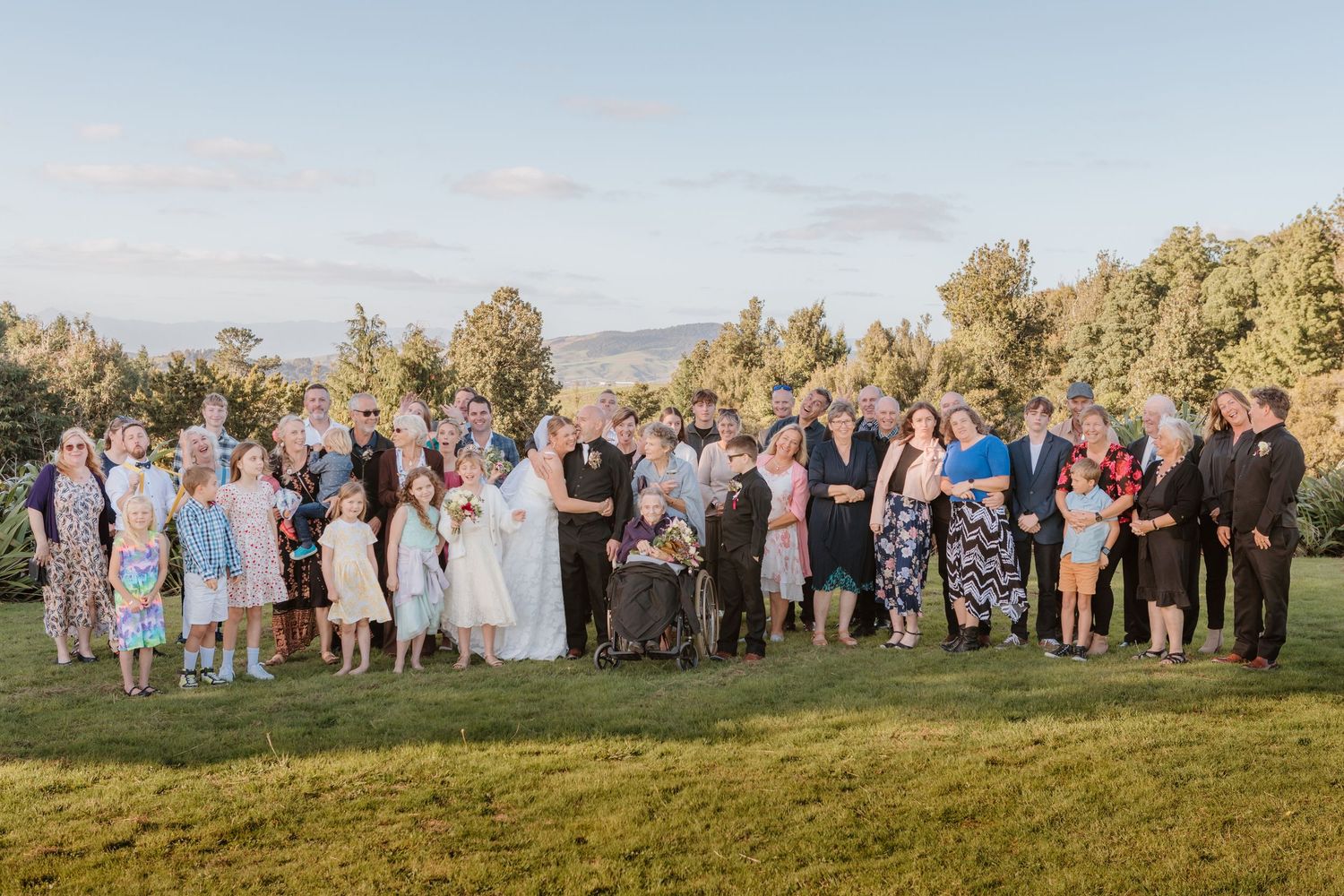 Large wedding party gathers for group photo on grassy hilltop during golden hour with mountains in background.