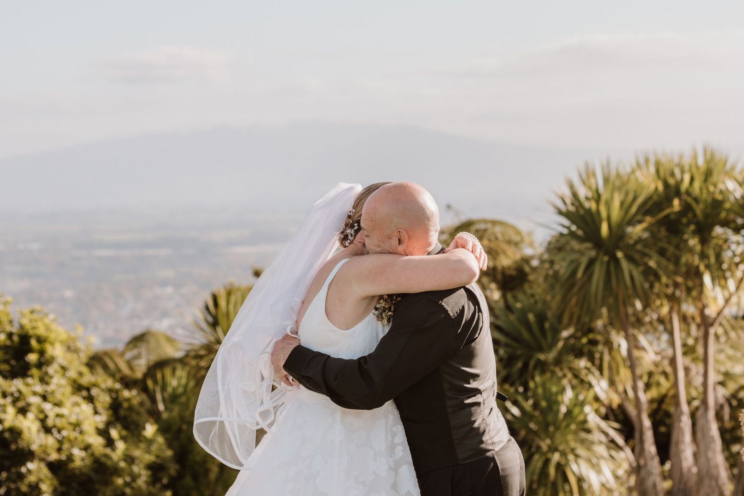 A couple embraces during their wedding ceremony at a scenic overlook with palm trees and ocean views at sunset.