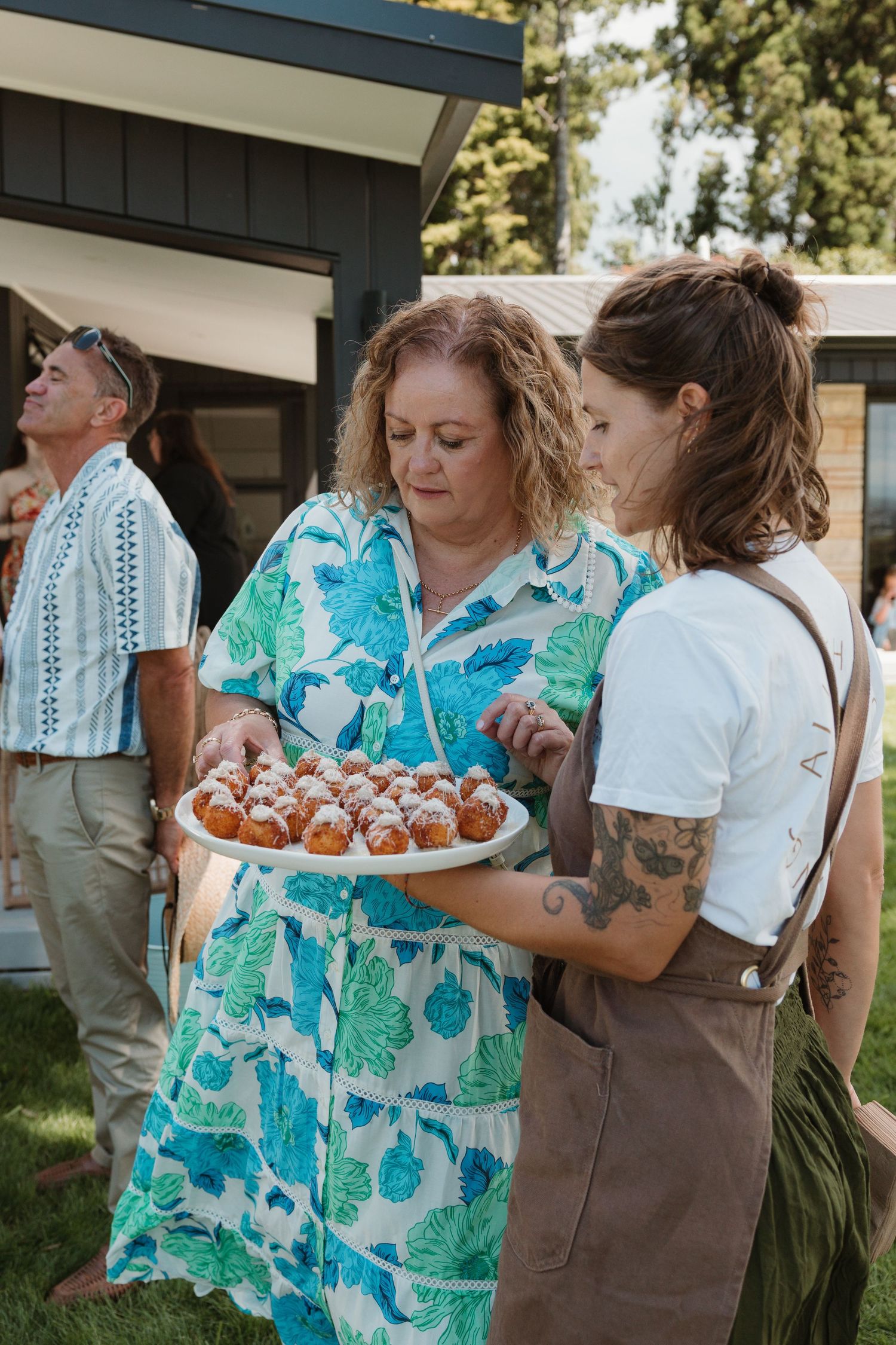 Two women admire a plate of powdered sugar pastries at an outdoor garden party event.