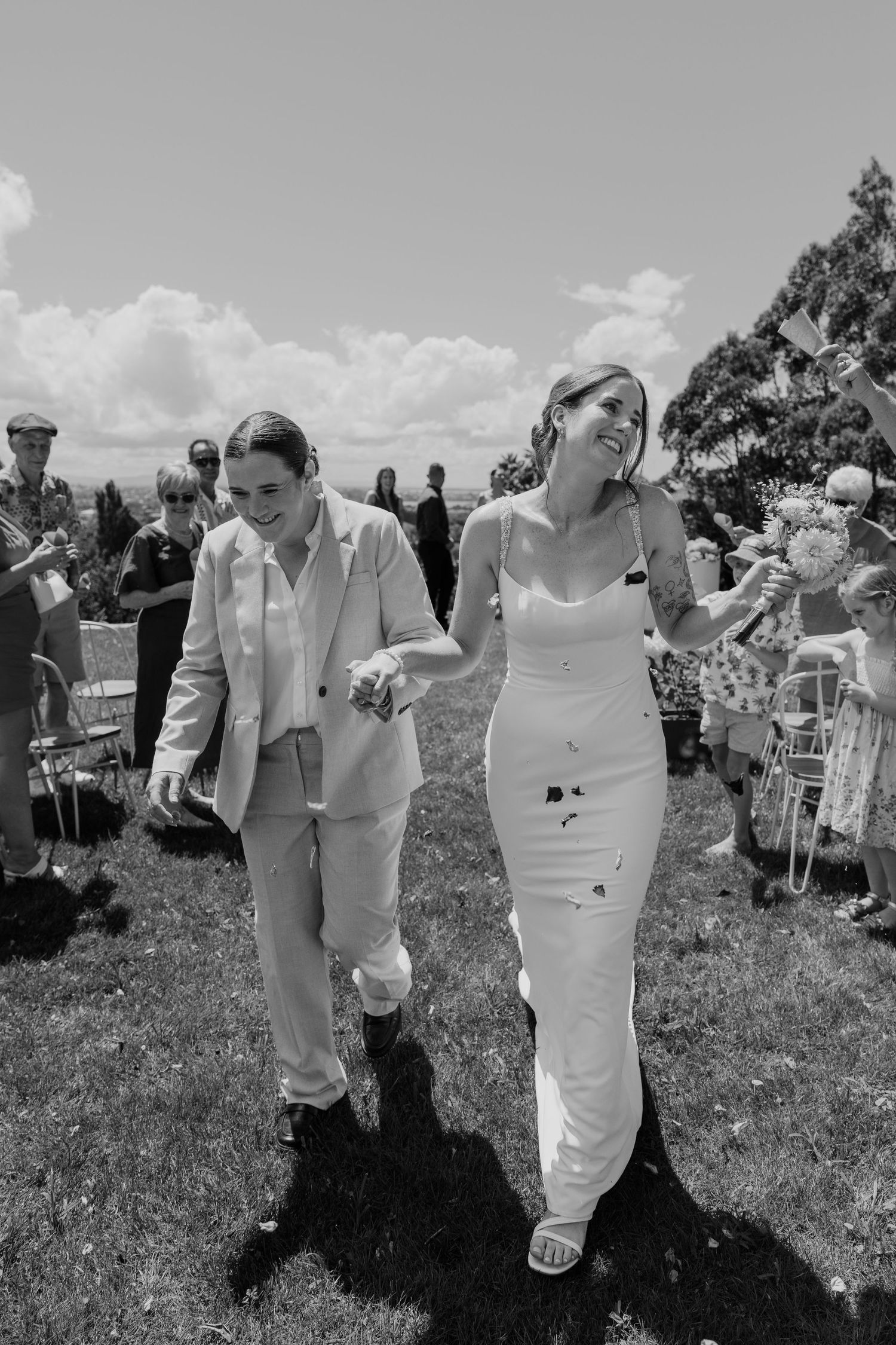 Black and white photo of two brides walking down the aisle, smiling, surrounded by guests at an outdoor wedding.