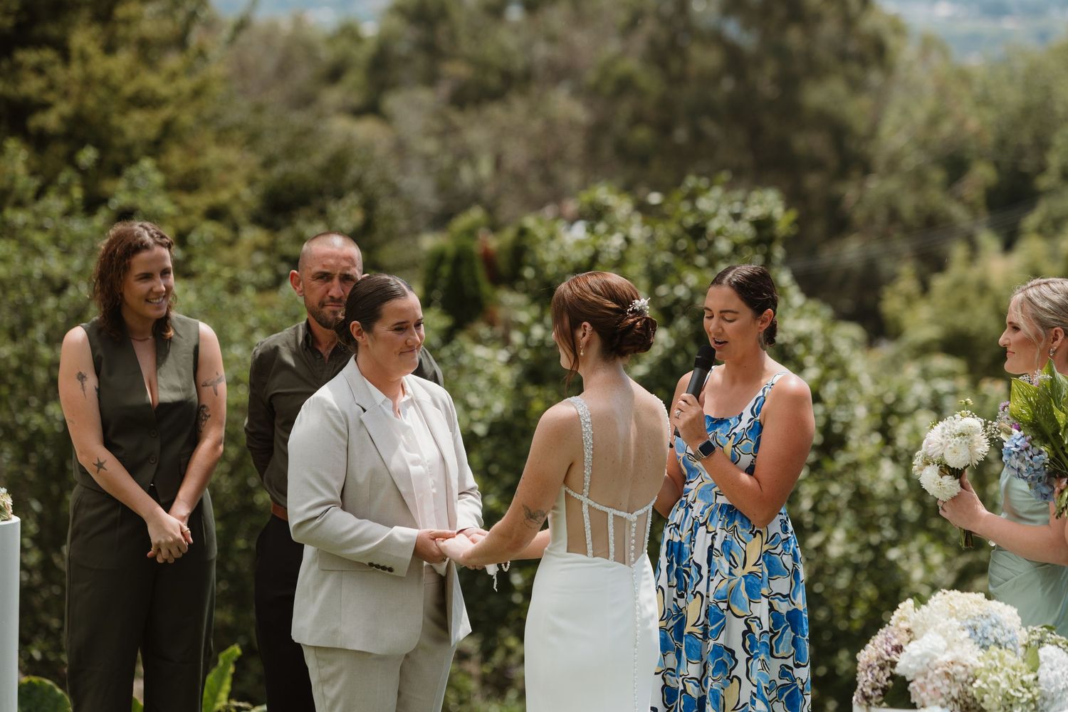 Two brides hold hands during an outdoor wedding ceremony surrounded by lush greenery and smiling guests.