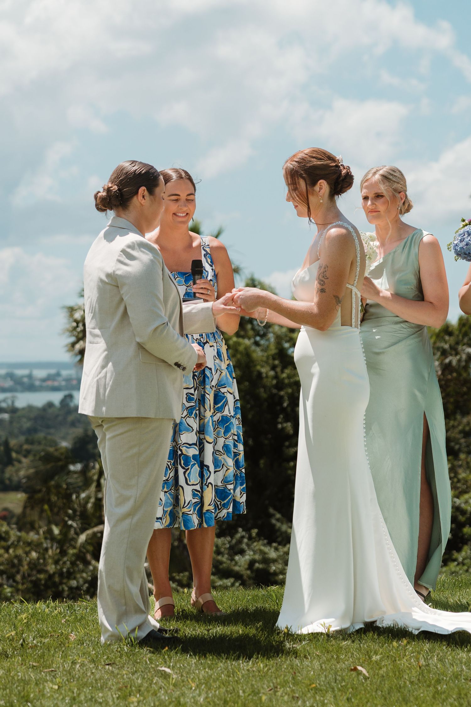 Bride and groom exchange rings outdoors on sunny day, with bridesmaid in mint gown and guest in blue floral dress.