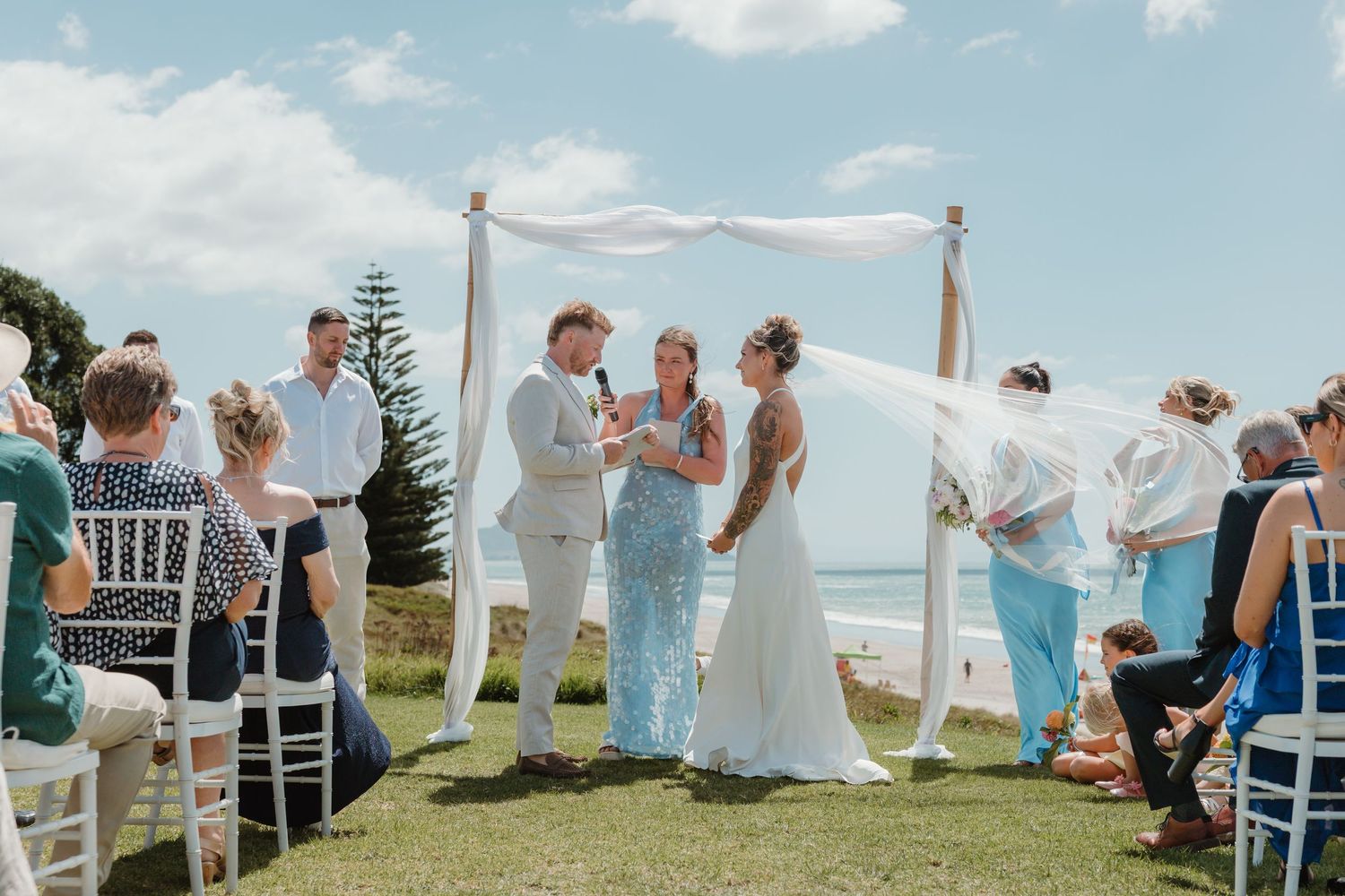 Outdoor beach wedding ceremony with couple exchanging vows under white draped arch, guests seated on white chairs.