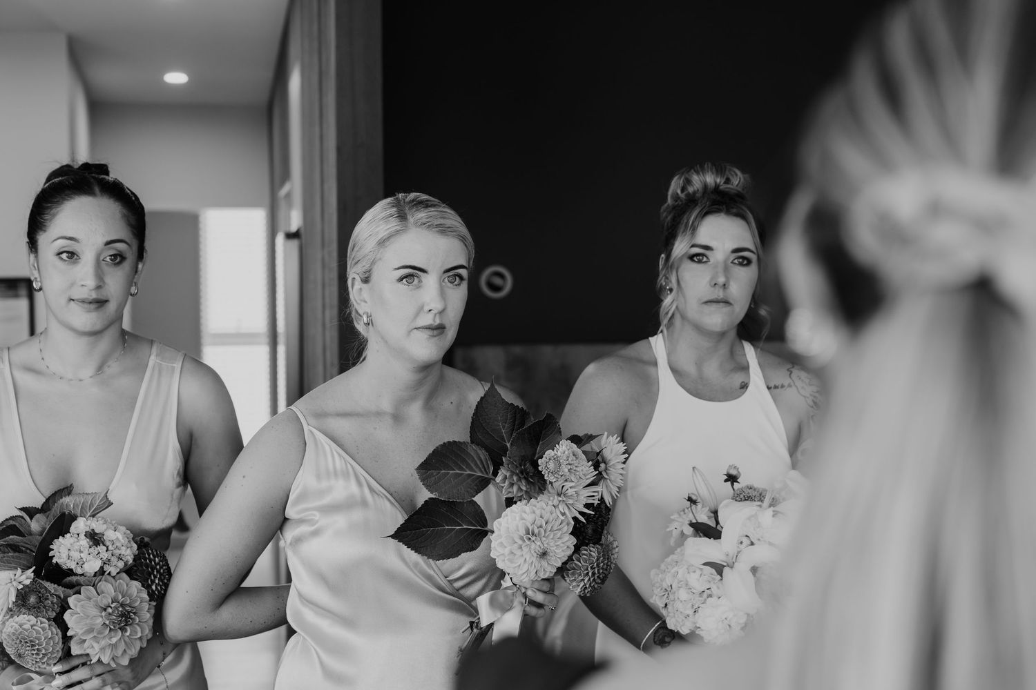 Three bridesmaids holding floral bouquets watch the bride in a candid black and white wedding preparation photo.