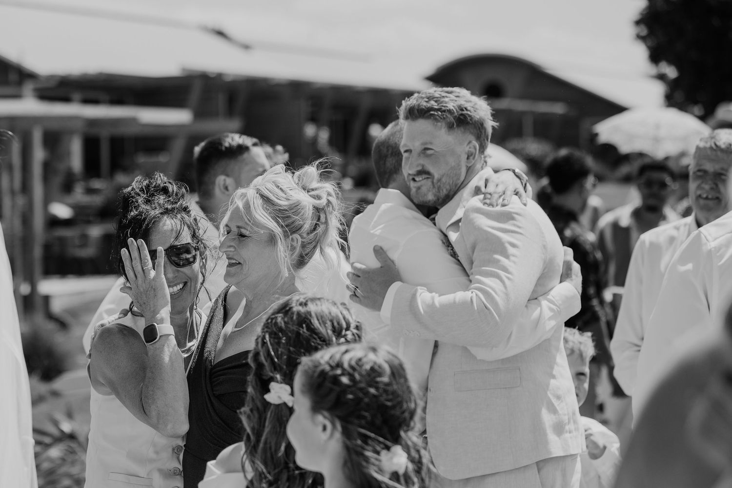 Black and white wedding photo of guests hugging and celebrating outdoors at a sunny reception.