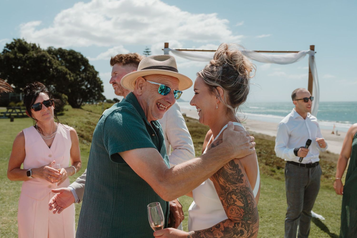 Couple embracing at outdoor beach wedding ceremony with guests in background on sunny day.