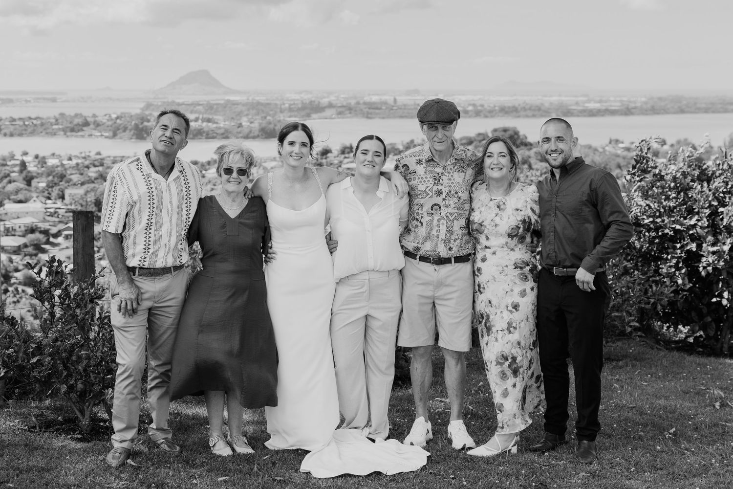 Black and white wedding photo of bride and groom with family group outdoors, scenic coastal view in background.