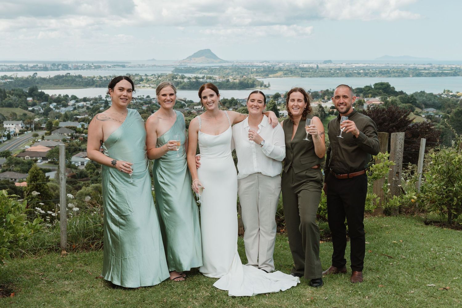 Bride with bridesmaids in mint green dresses and family posing on a scenic hilltop overlooking a bay and mountain.