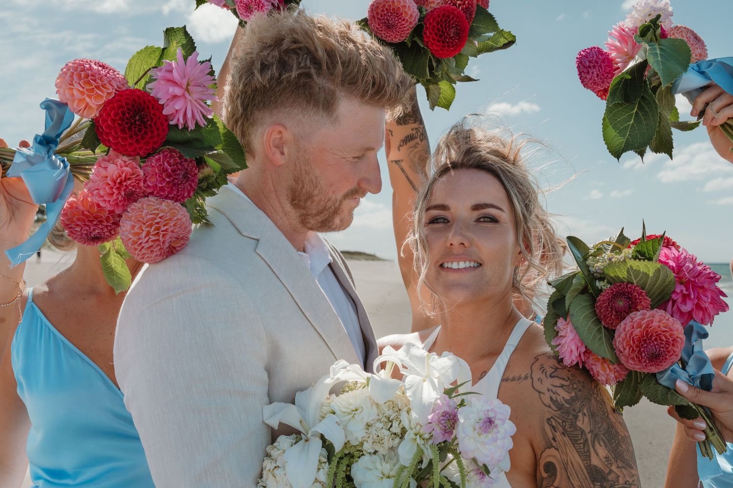 Couple exchanging vows at outdoor beach wedding ceremony under floral arch with pink dahlias and blue bridesmaid.