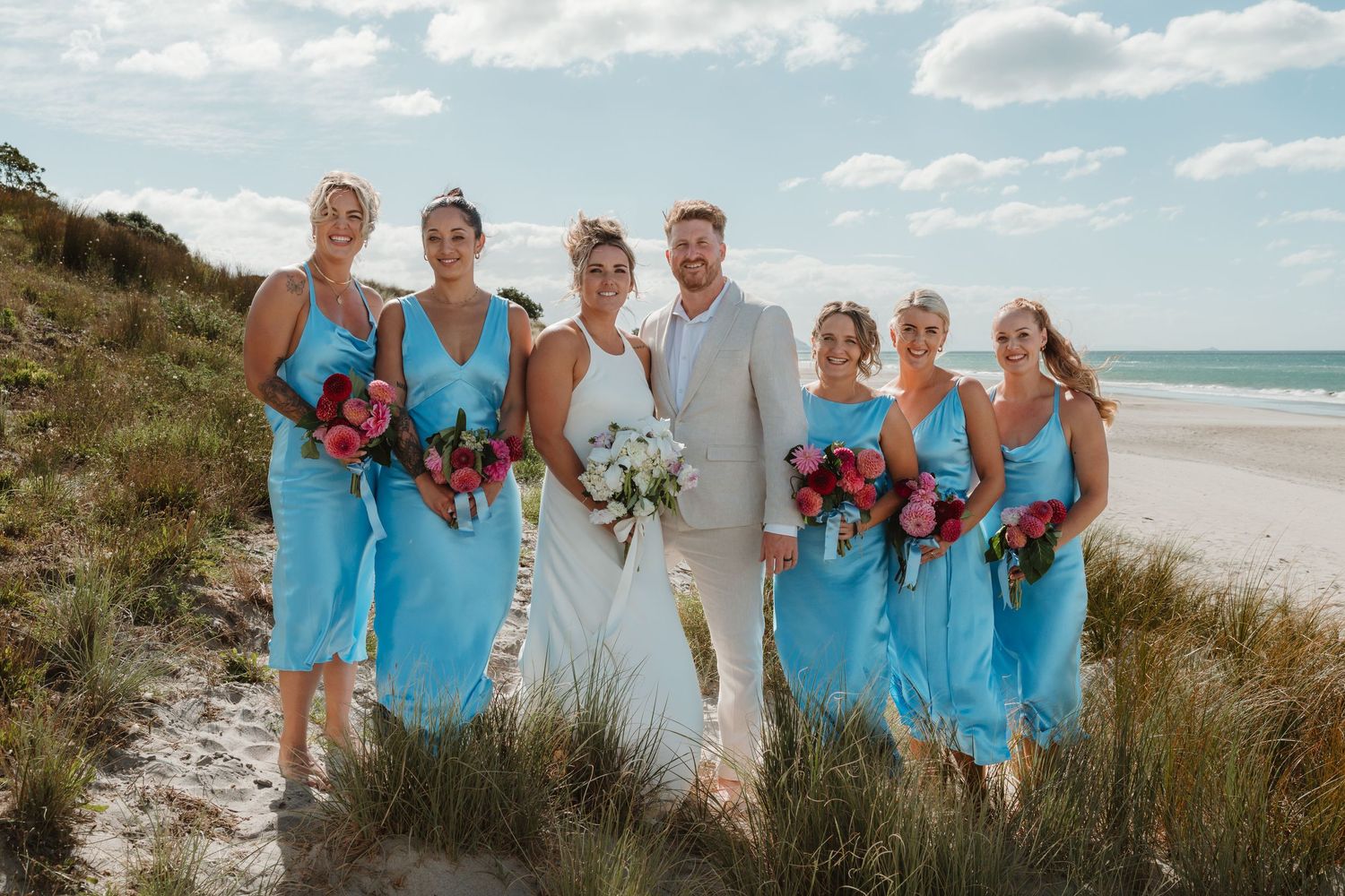 Bride and groom with four bridesmaids in blue dresses holding pink bouquets on a sandy beach.