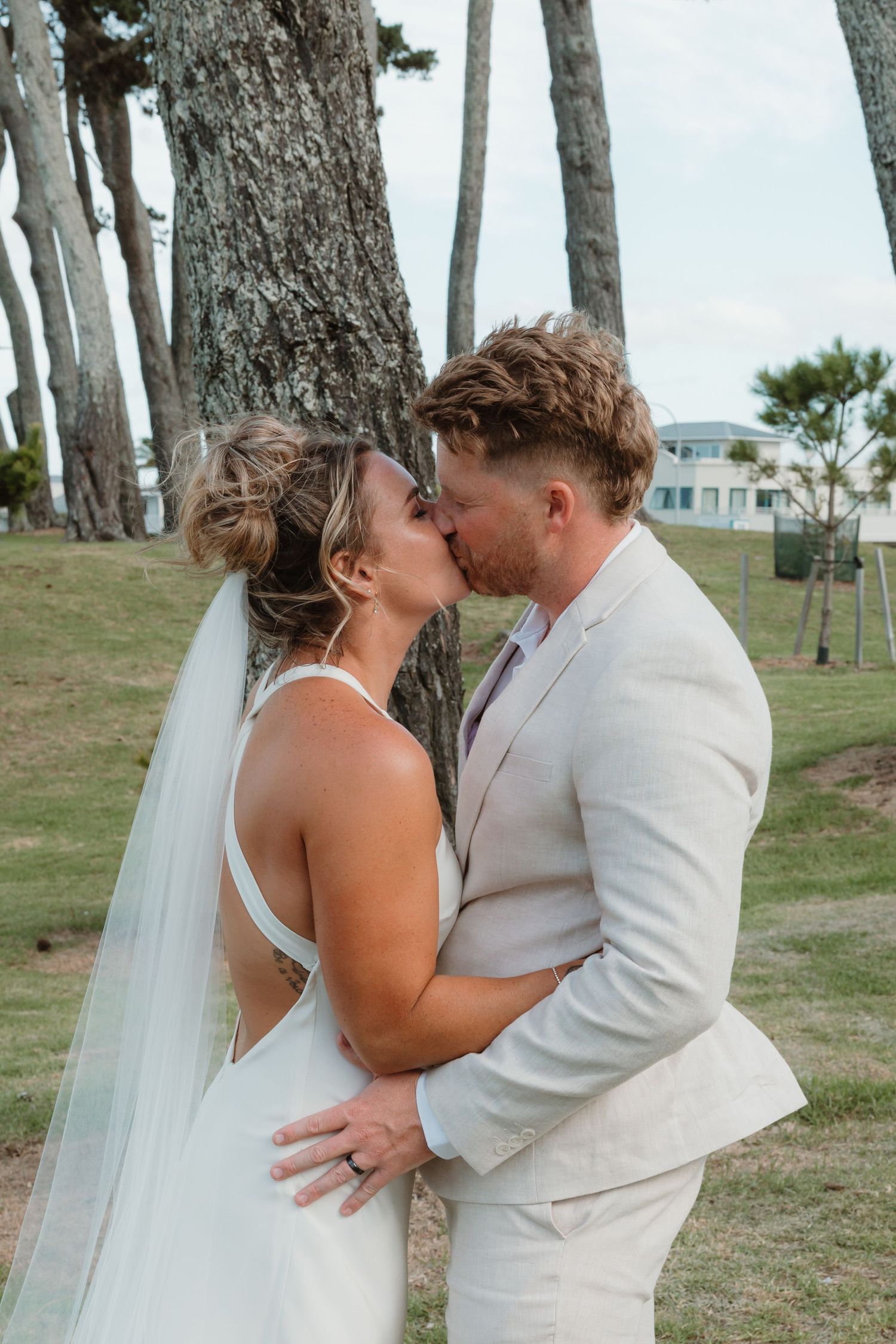 A bride and groom share a romantic kiss outdoors near tall trees, dressed in a white gown with veil and beige suit.