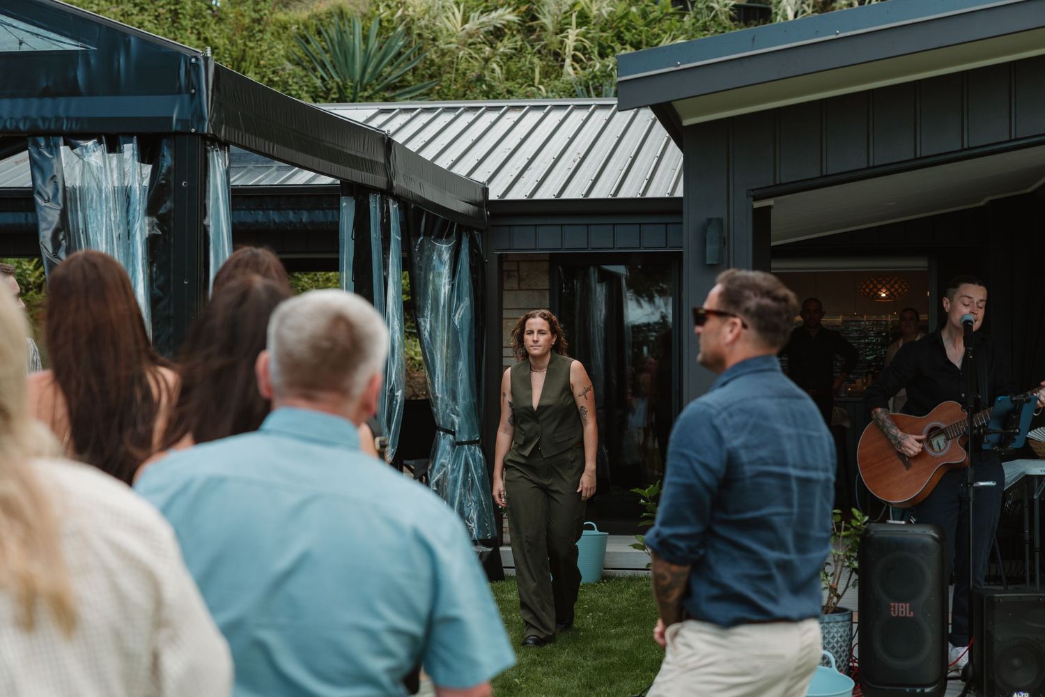 Outdoor gathering with guests watching a performer walk toward them as a guitarist plays live music near a modern home.