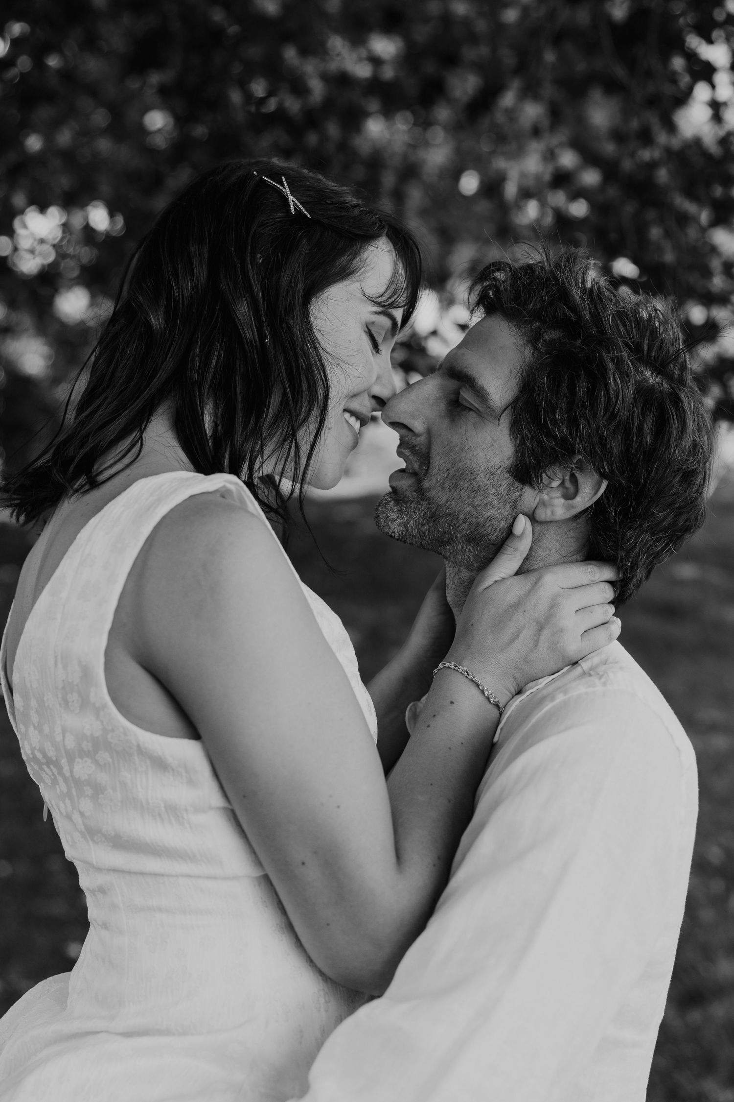 Black and white photo of a romantic couple about to kiss outdoors, woman in white dress tenderly holding mans face.