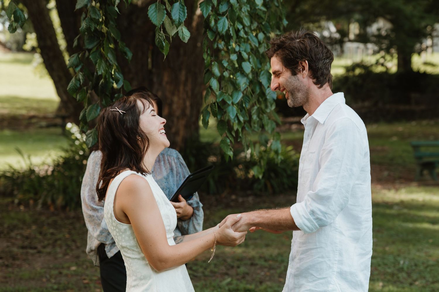 Couple laughing and holding hands during an outdoor wedding ceremony surrounded by lush green trees.