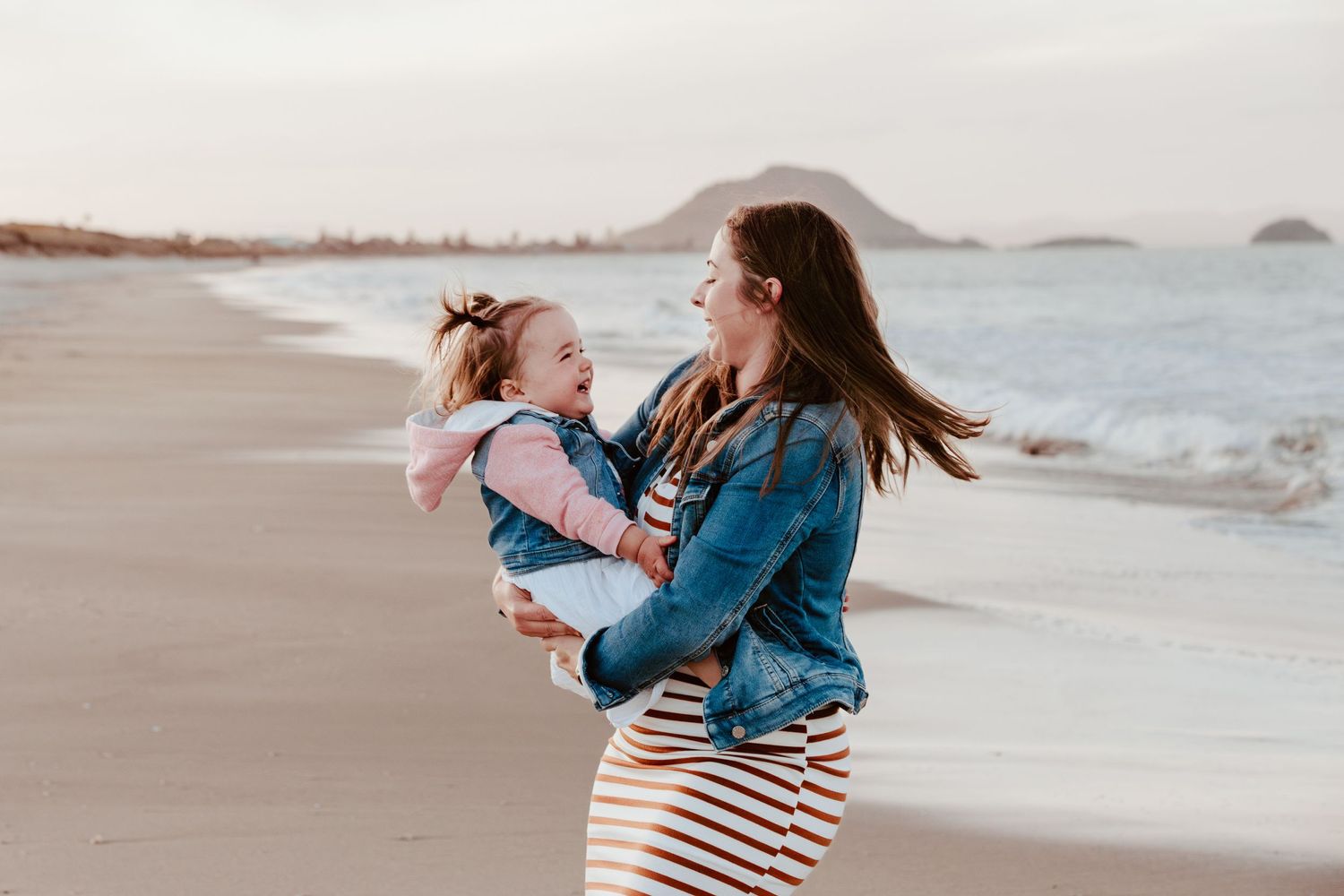Mum and daughter share a joyful moment on a serene beach with Mount Maunganui visible in the background at sunset. Tauranga photographer