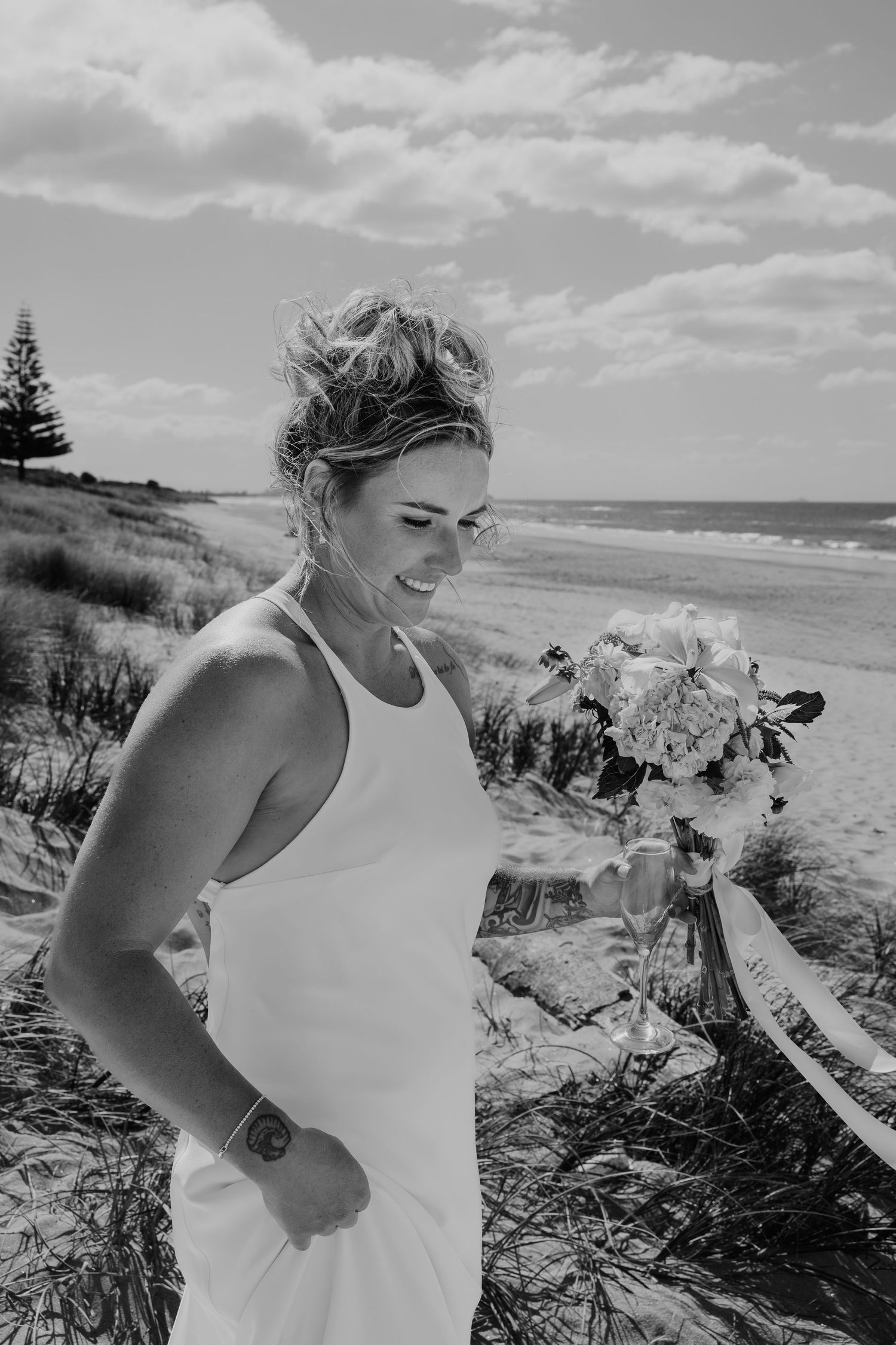 Black and white photo of a smiling bride in a white dress holding a bouquet on a sandy beach.