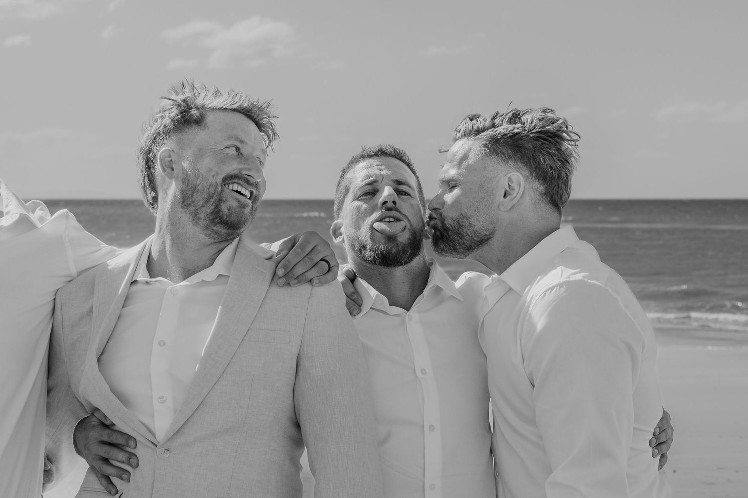 Three men in white suits laugh and share a joyful moment together on a beach at a wedding celebration.