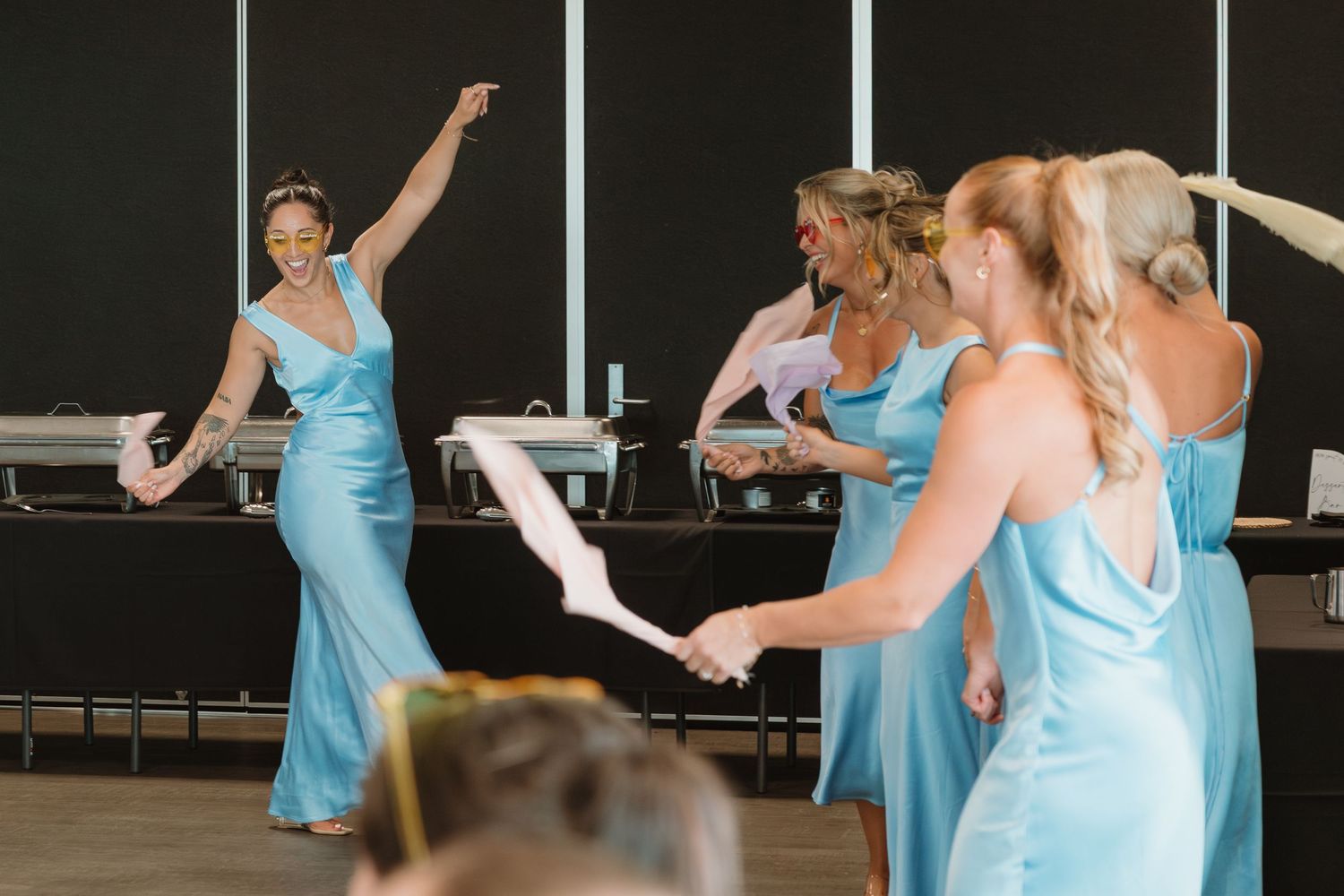 Bridesmaids in light blue satin gowns dancing and celebrating at a wedding reception near catering tables.