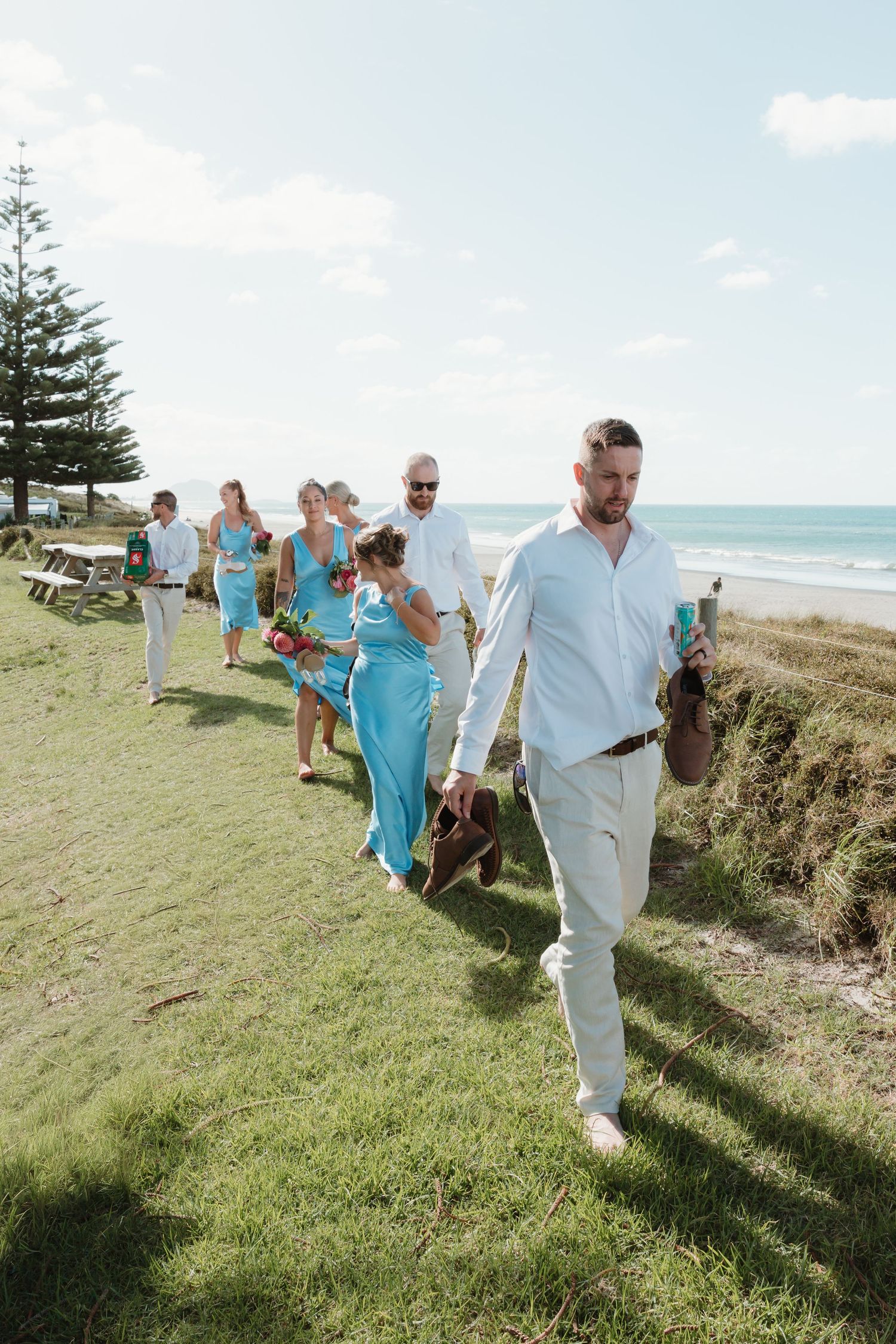 Wedding party in turquoise attire walks along grassy beachside path on sunny day.