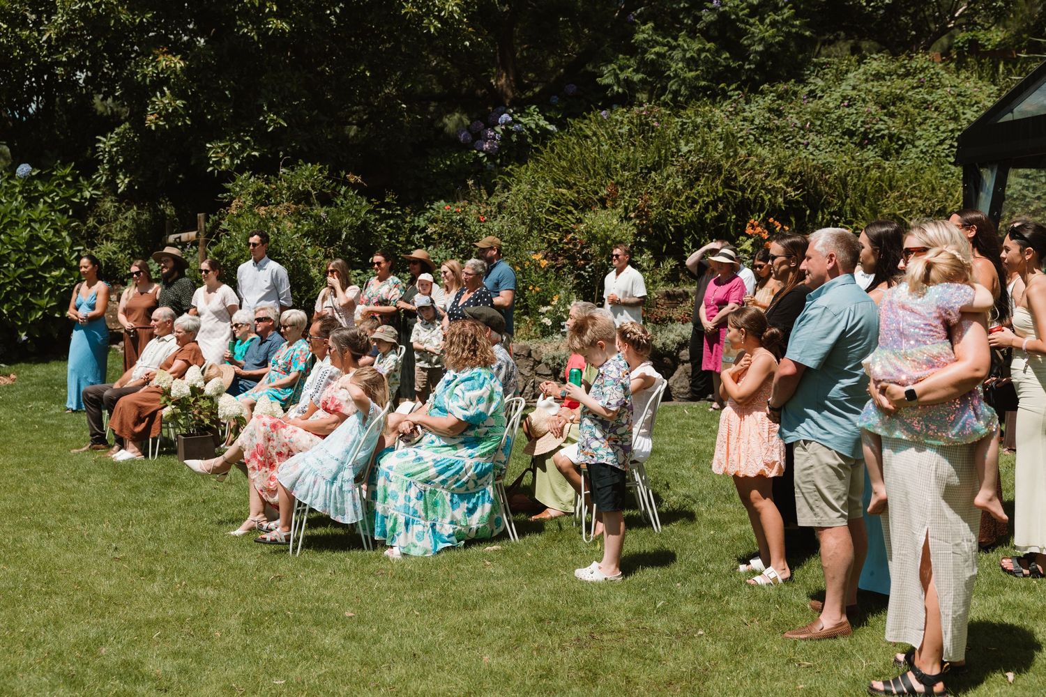 Guests gathered outdoors on sunny lawn for elegant garden wedding ceremony, seated in chairs surrounded by lush green trees.