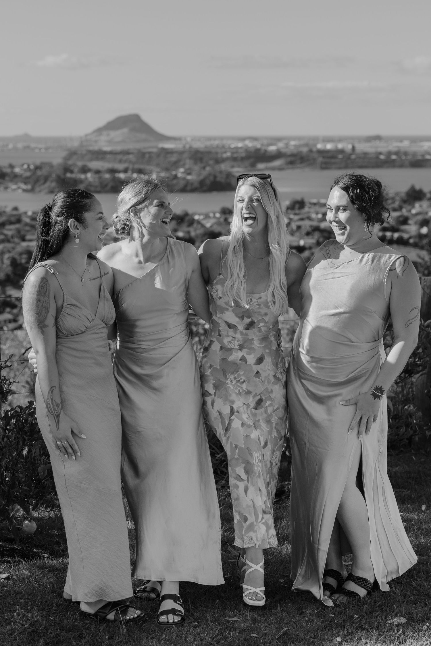 Four women in elegant gowns laugh together outdoors with a scenic coastal hill backdrop in black and white.