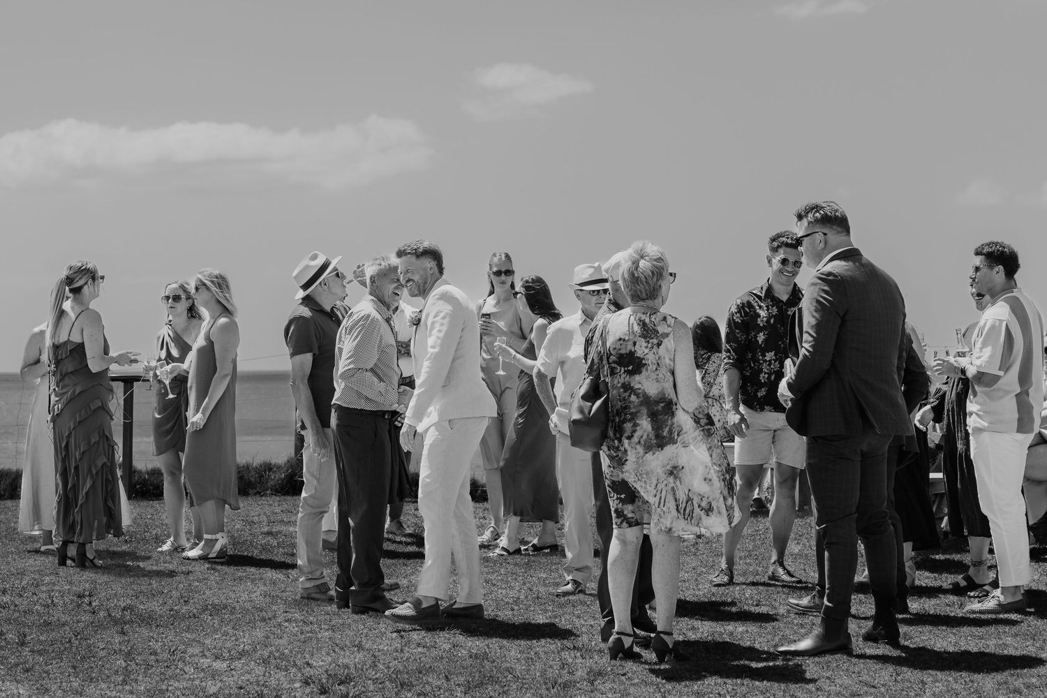 Black and white photo of elegantly dressed guests gathered outdoors at a seaside wedding reception.