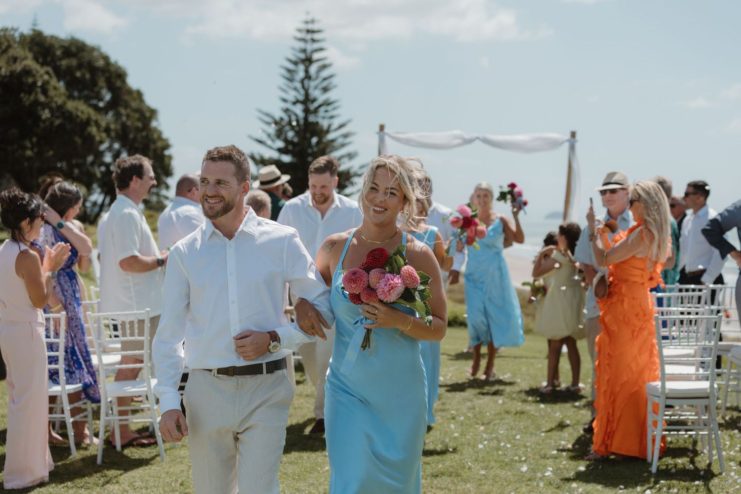 Couple walks down outdoor wedding aisle on grassy coastal venue, guests applauding, bridesmaid in blue dress holding pink bouquet.