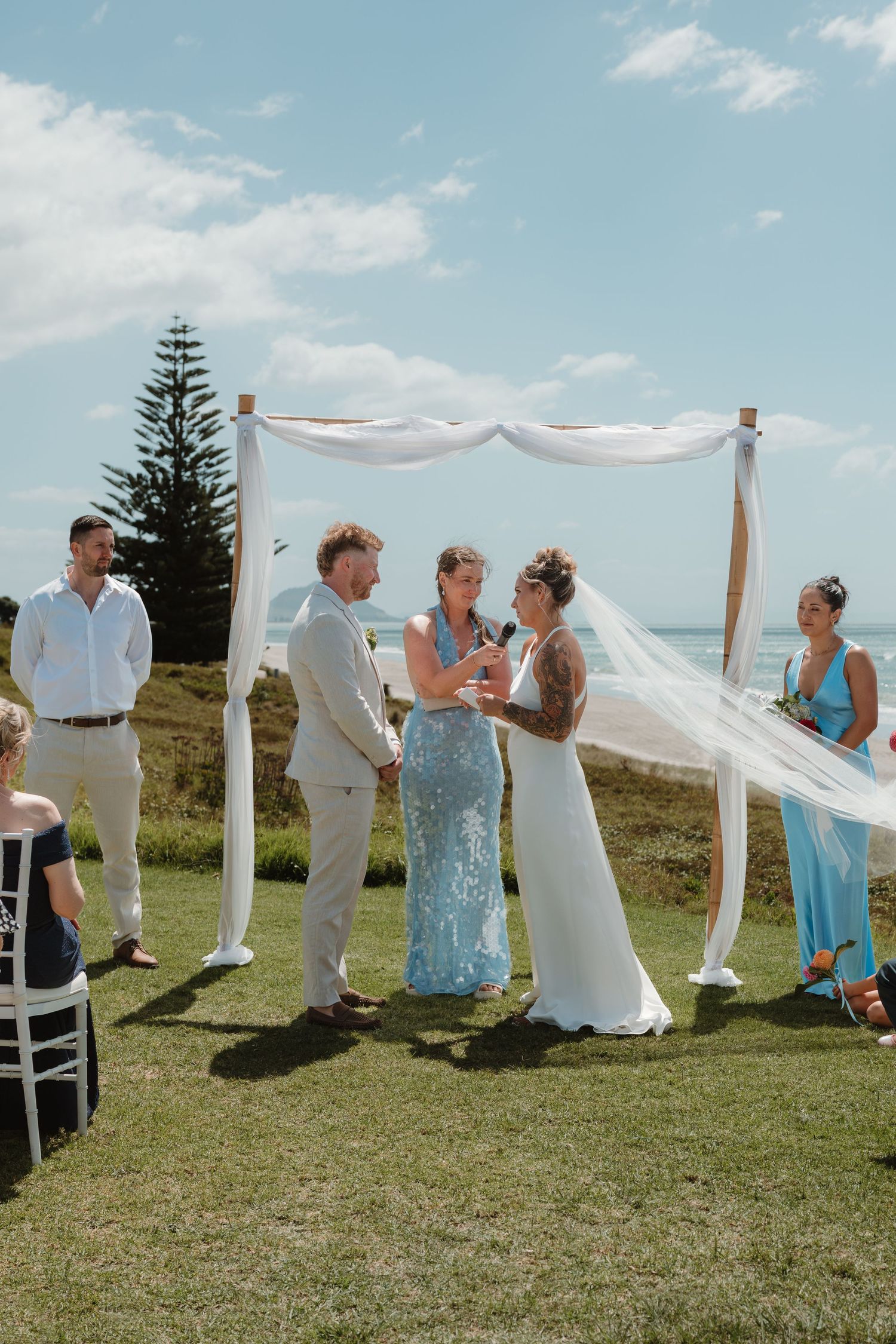 Bride and groom holding hands at an outdoor coastal wedding ceremony under a white draped arch.