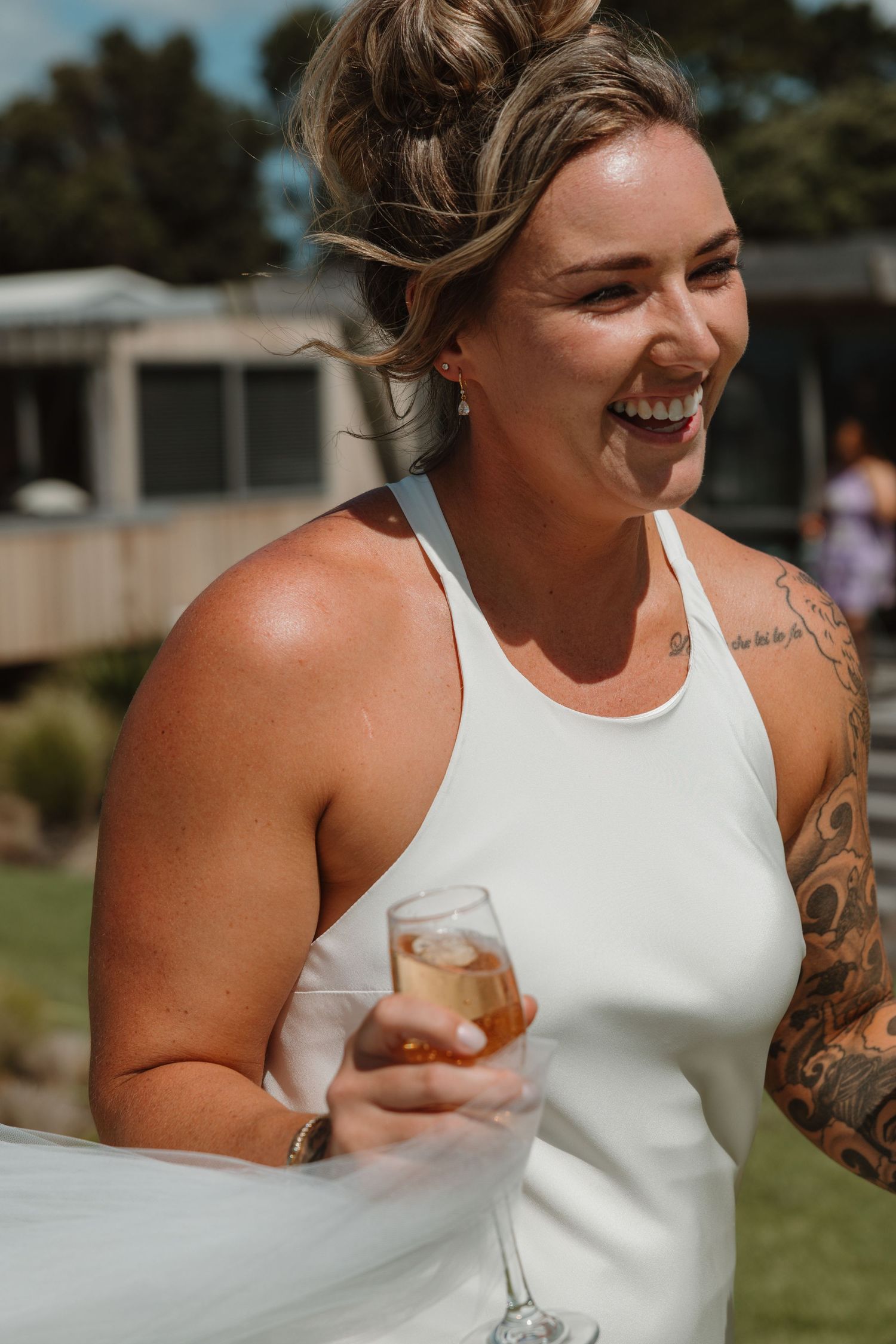 Laughing bride in white dress holding champagne glass outdoors, showing tattooed arm at wedding celebration.