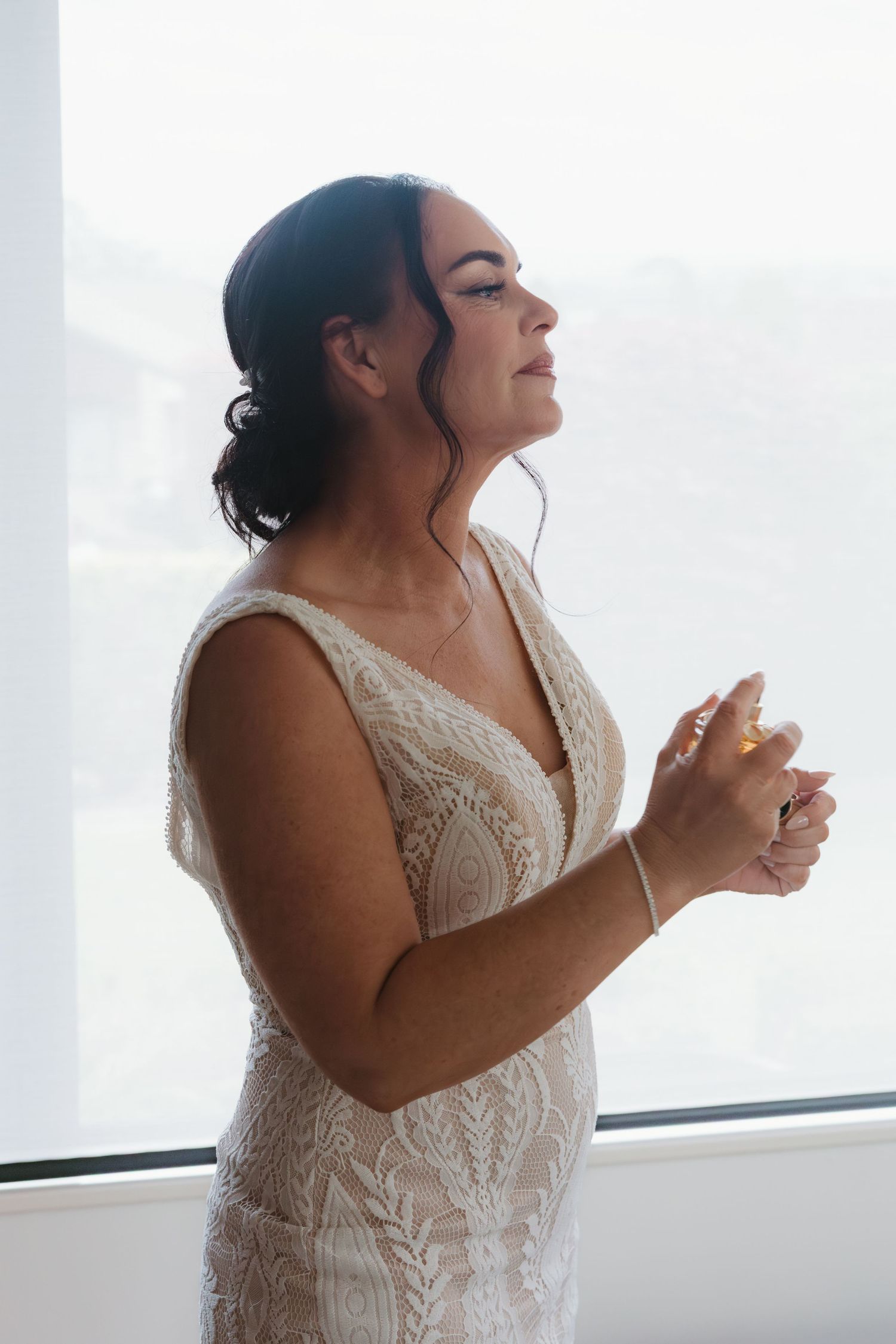 A person in a white lace wedding dress laughs joyfully while getting ready by a bright window.