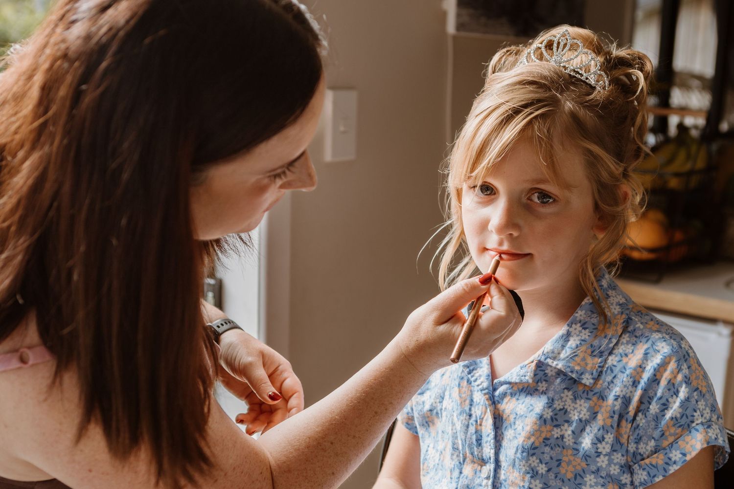 A person applies makeup on a young child wearing a blue floral shirt and sparkly tiara in warm indoor lighting.
