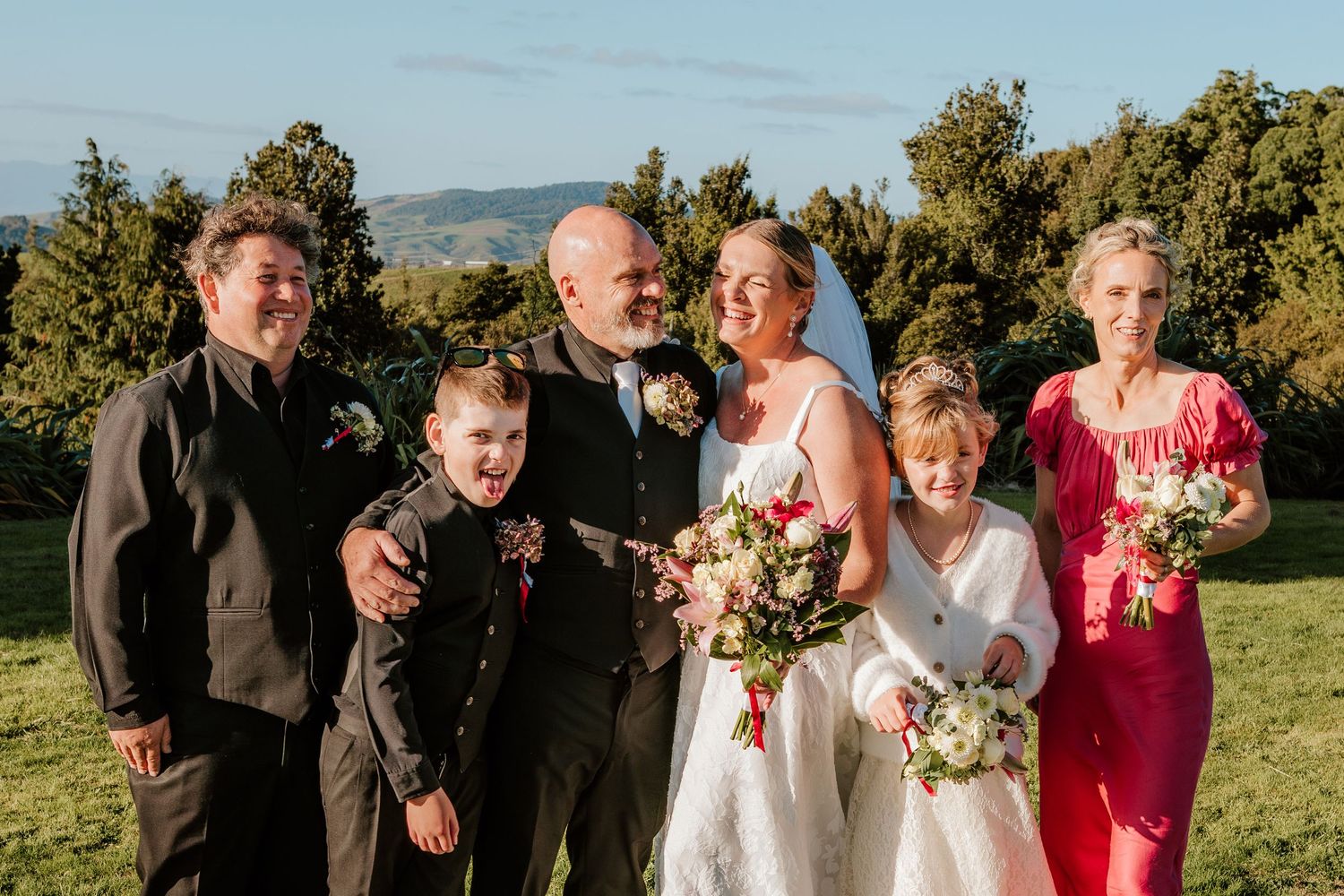 A wedding party poses together in a scenic outdoor setting with mountains and trees in the background.