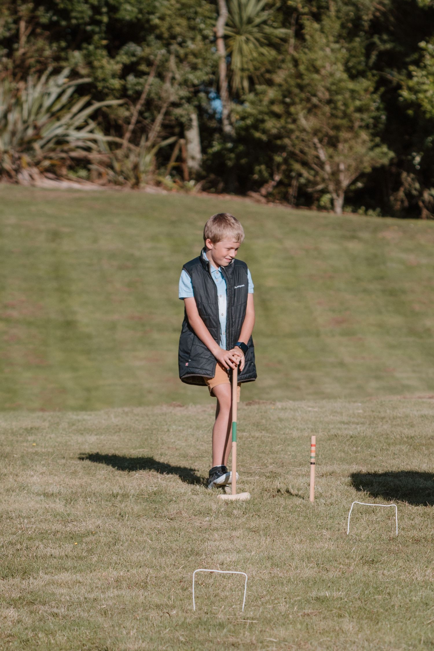 Young player in a blue shirt and dark vest concentrates while playing croquet on a sunny grassy lawn.