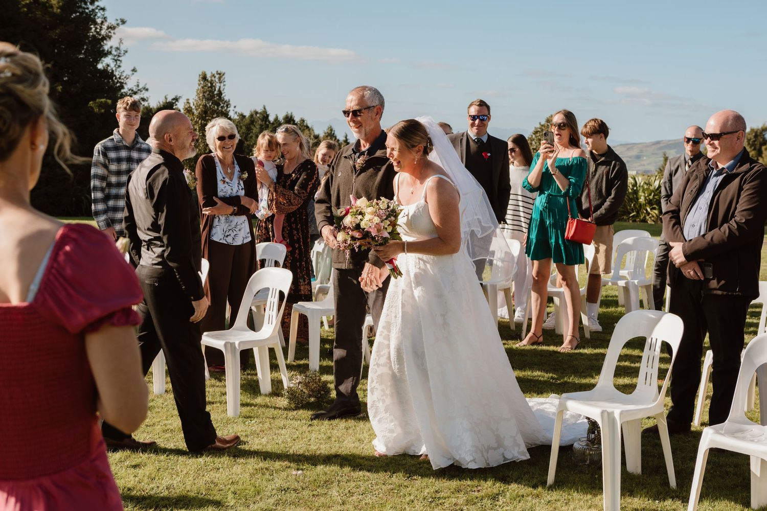 A sunlit outdoor wedding ceremony on a grassy hill with white chairs and guests gathered around.