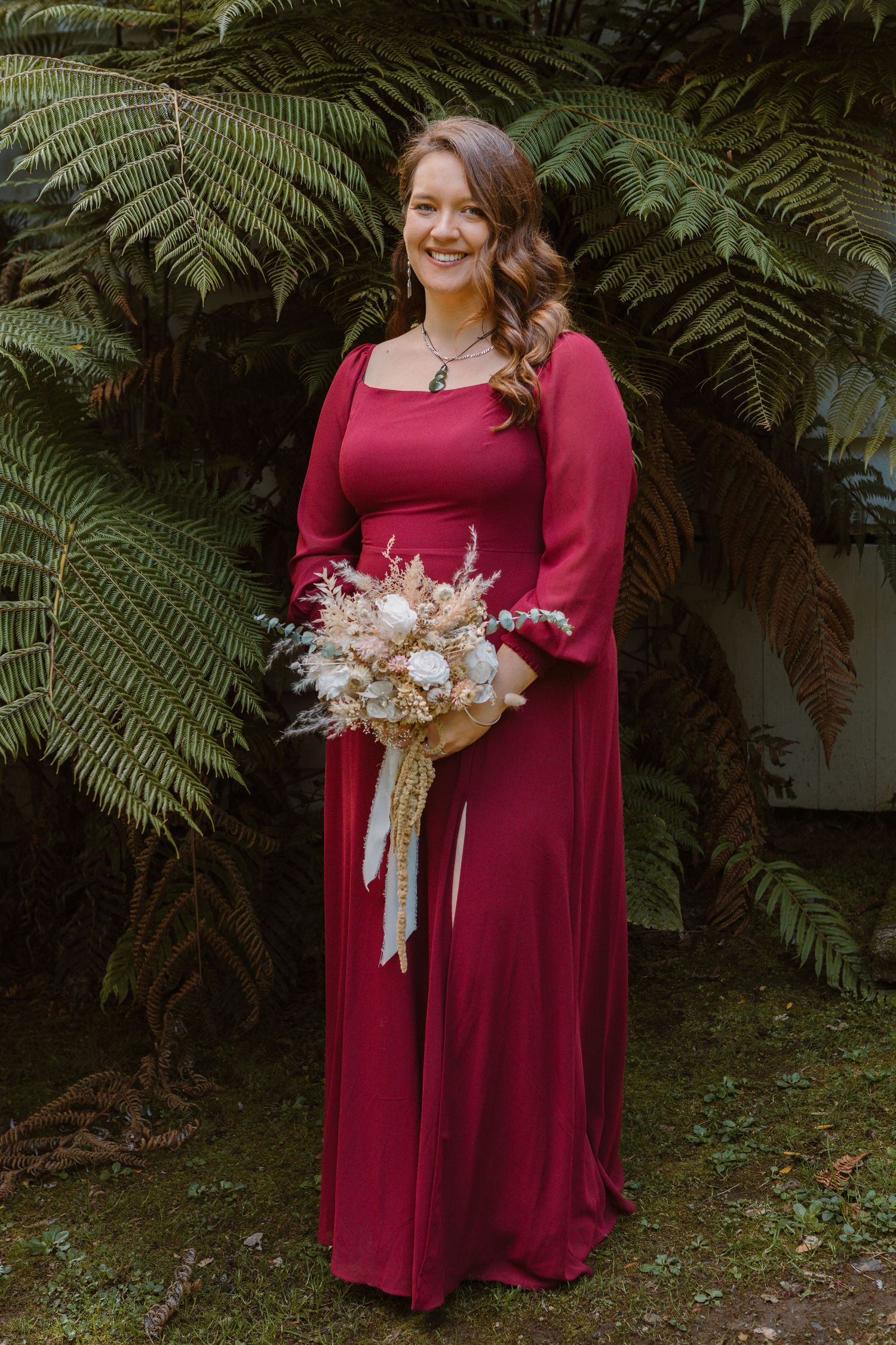 Someone in a burgundy long sleeve formal dress holds a dried flower bouquet while standing in front of large ferns.