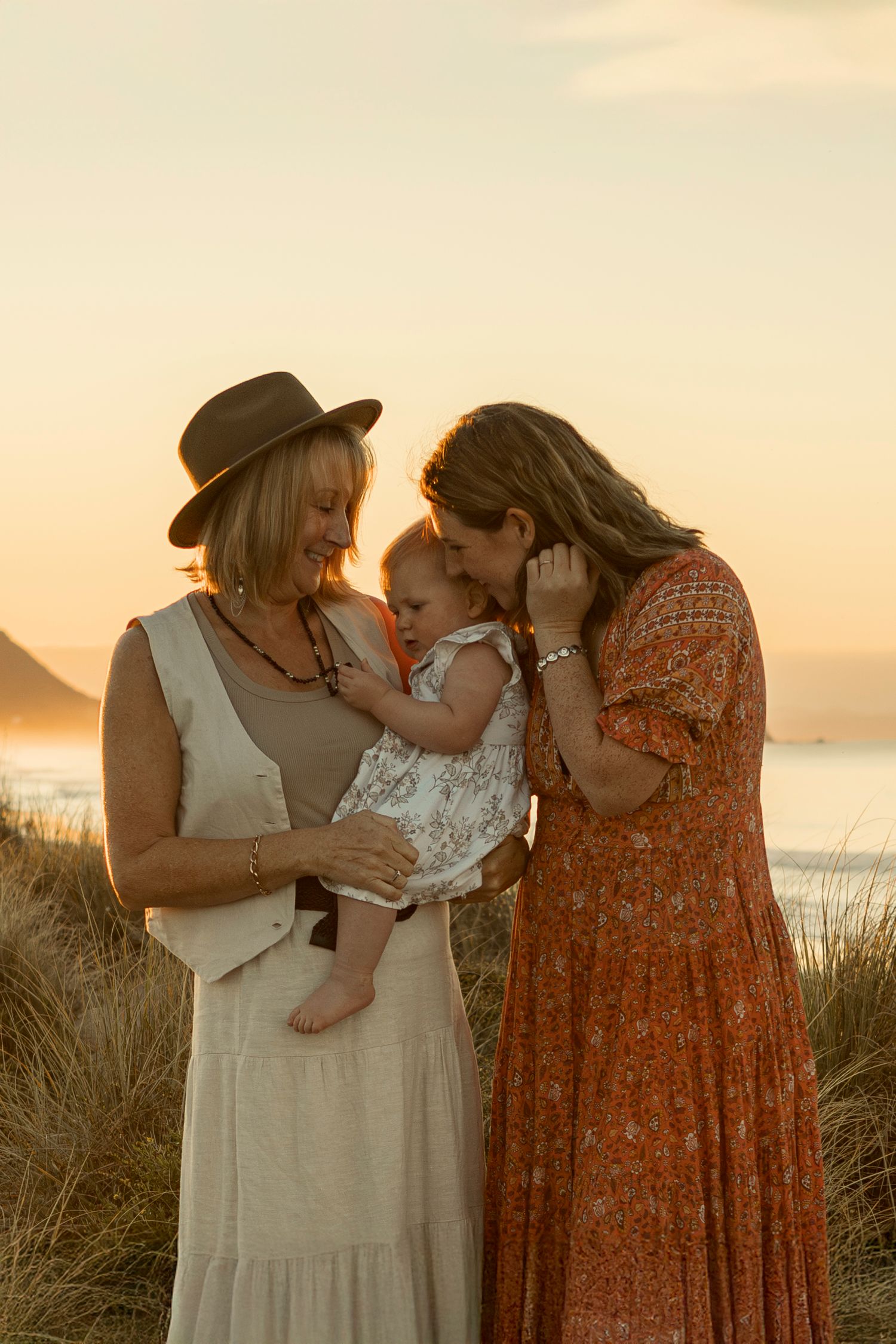 Three figures share a tender moment at sunset on a beach with tall grass and golden light in the background.