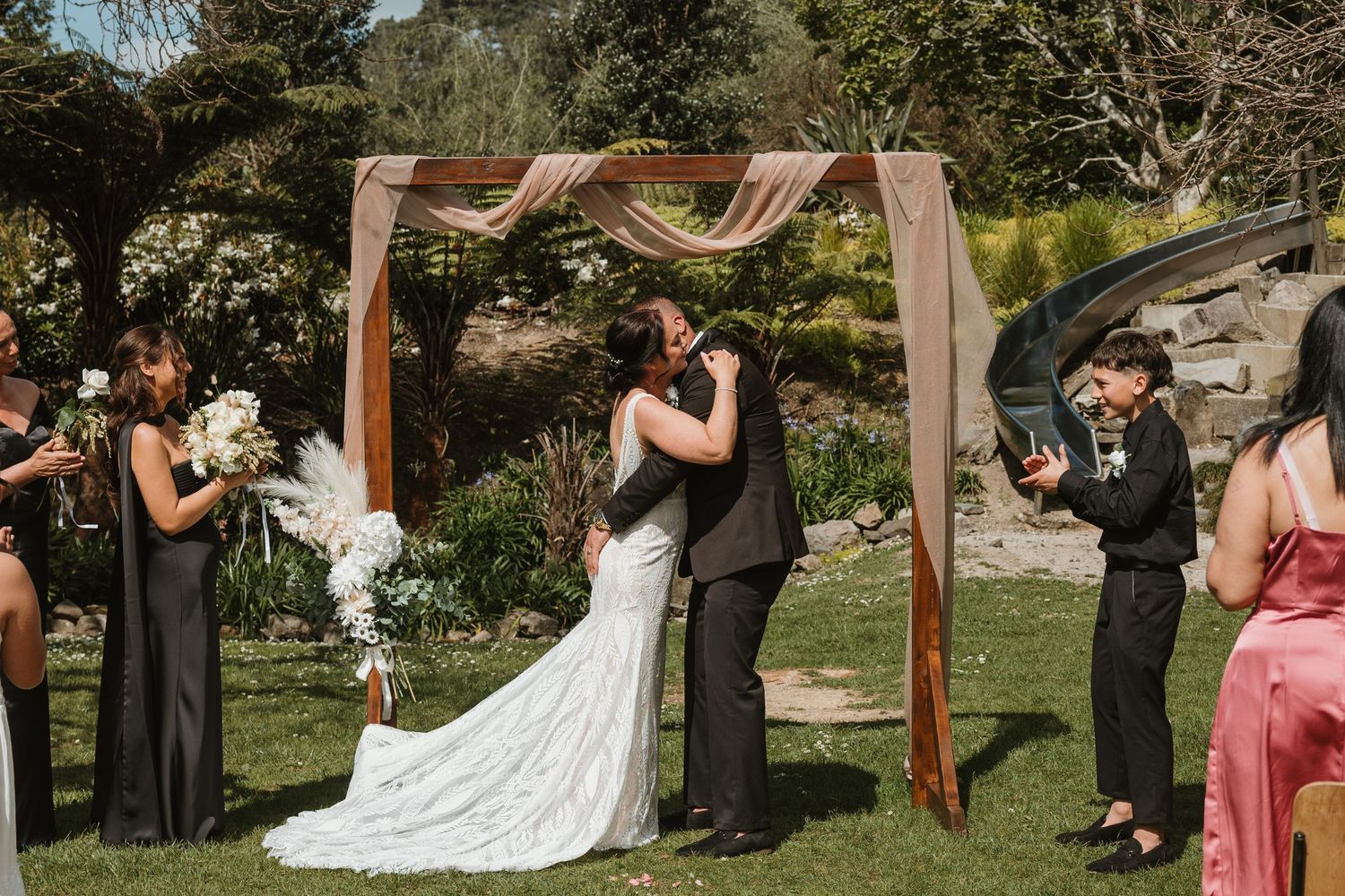 A wedding ceremony takes place under a wooden arch in a sunny garden setting with guests looking on.