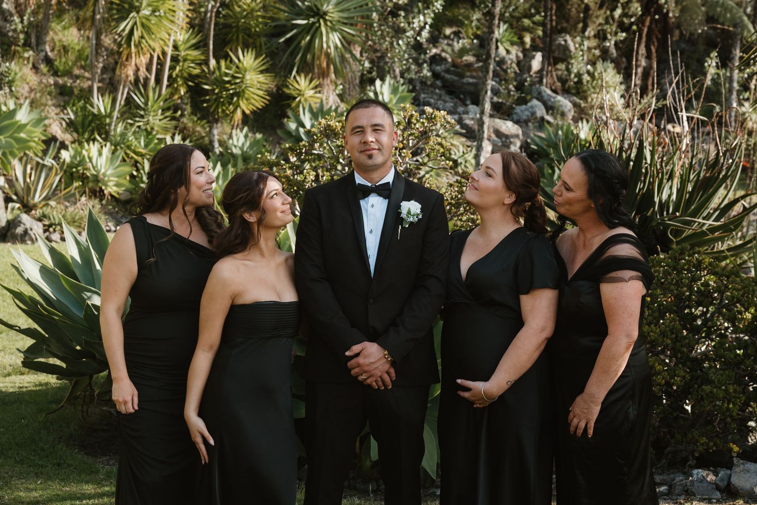 A group in formal black attire poses together in a lush garden setting with tropical plants and palm fronds.